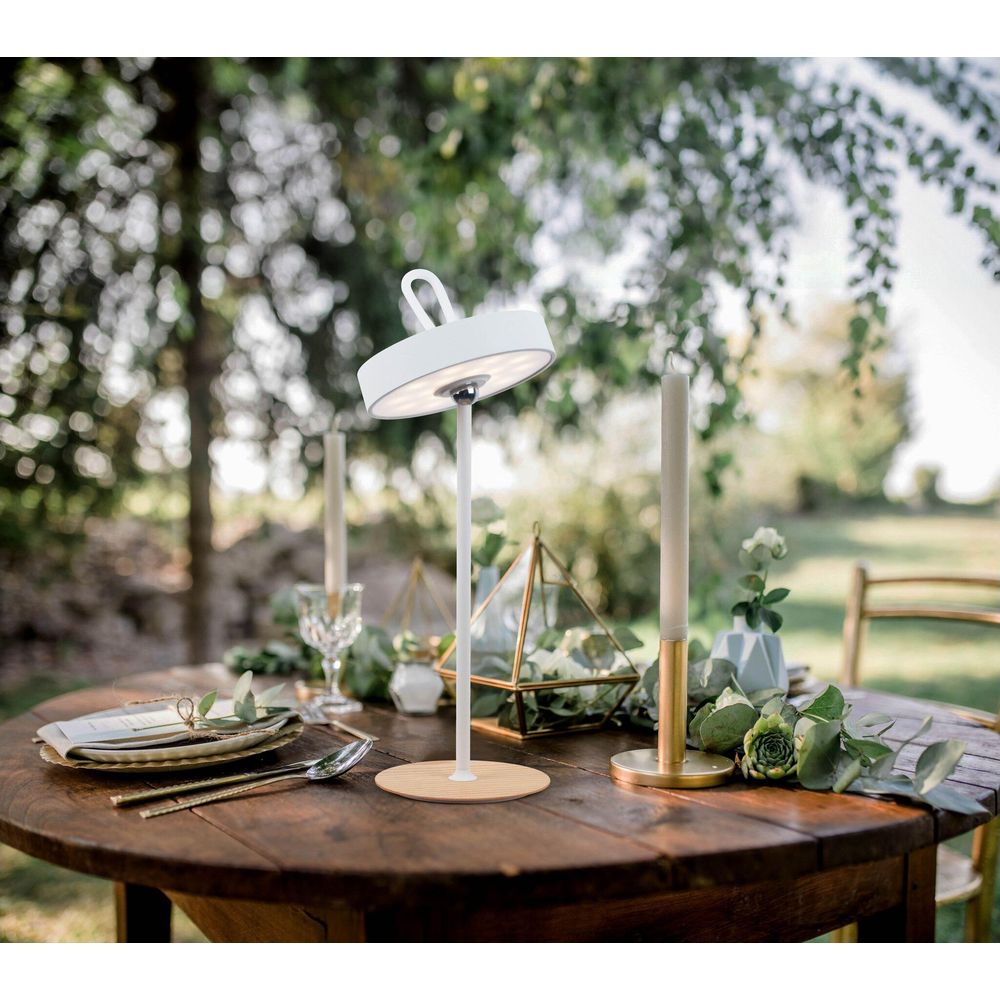 Weiße Tischlampe auf einem Holztisch mit Kerzen und Pflanzen im Freien.