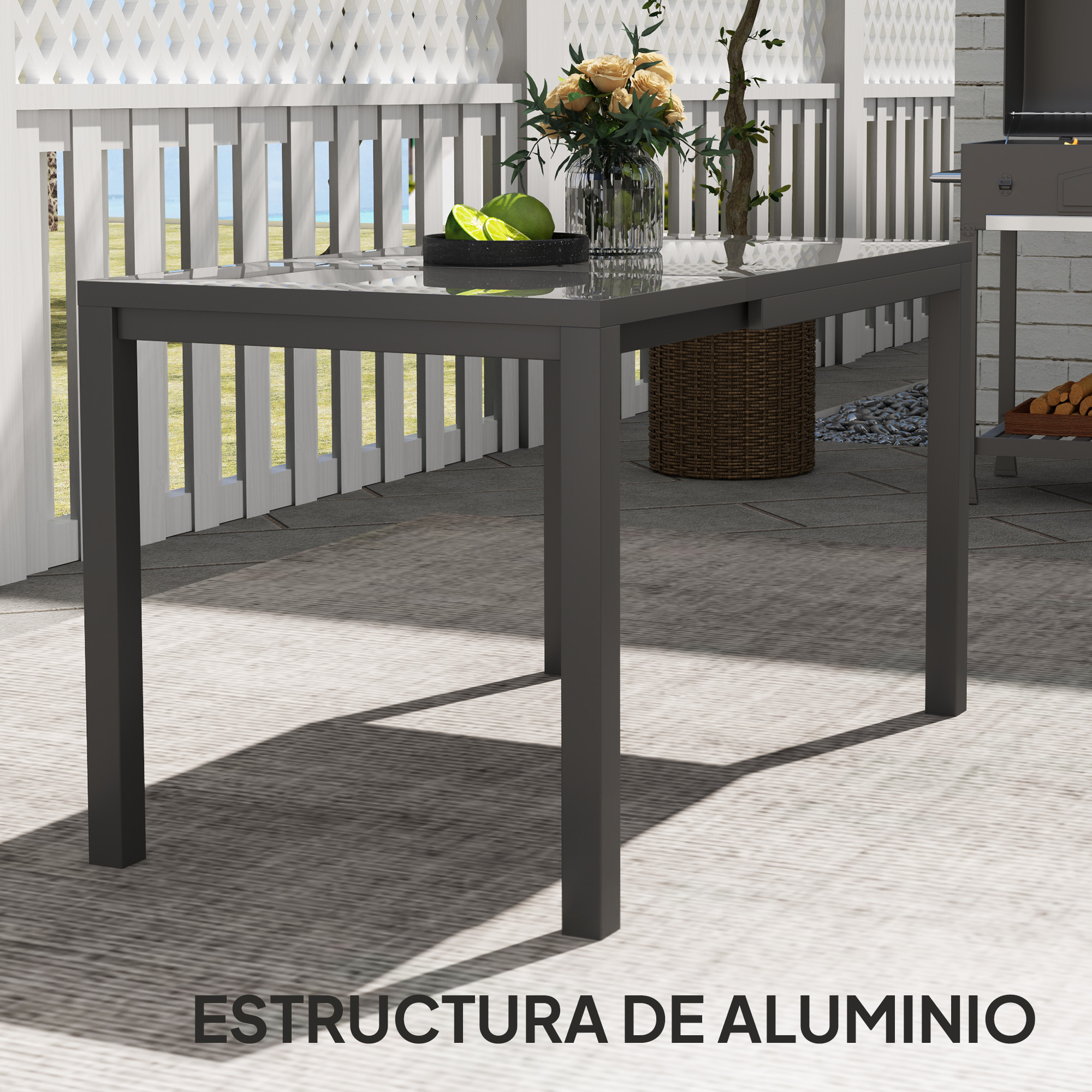 Una mesa gris con tapa de cristal en un patio. Tiene flores y limas encima.