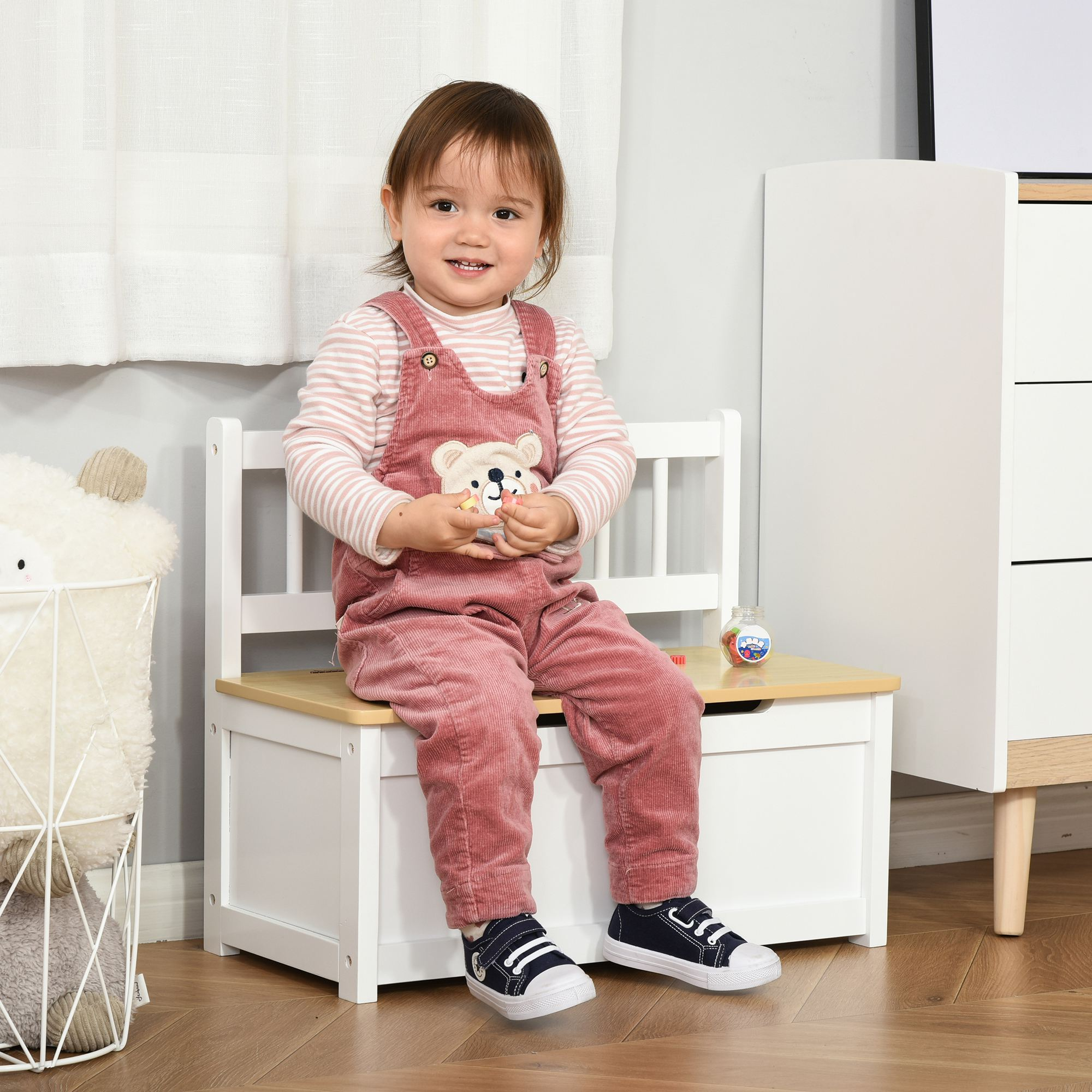 Un niño pequeño se sienta en un banco blanco con caja de juguetes, sosteniendo juguetes.