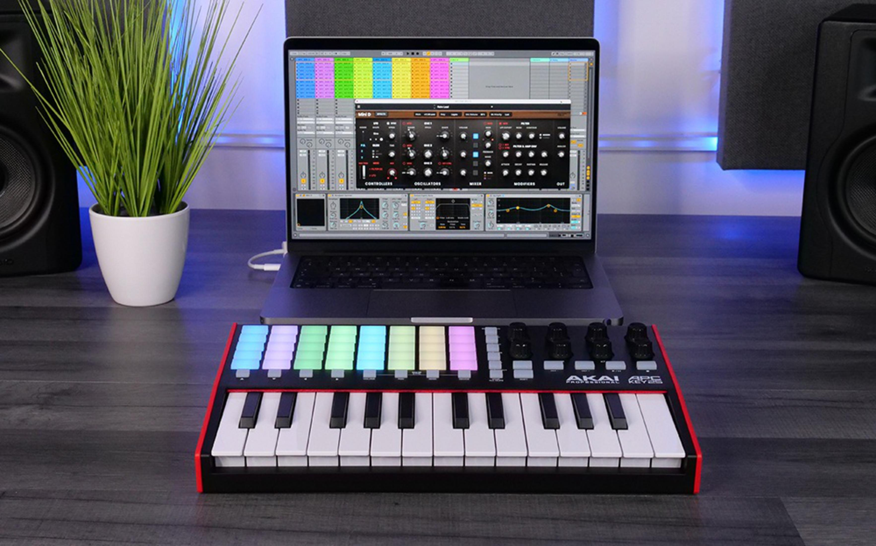 Laptop z oprogramowaniem muzycznym stoi za mini-klawiaturą. Po lewej stronie znajduje się zielona roślina w doniczce.