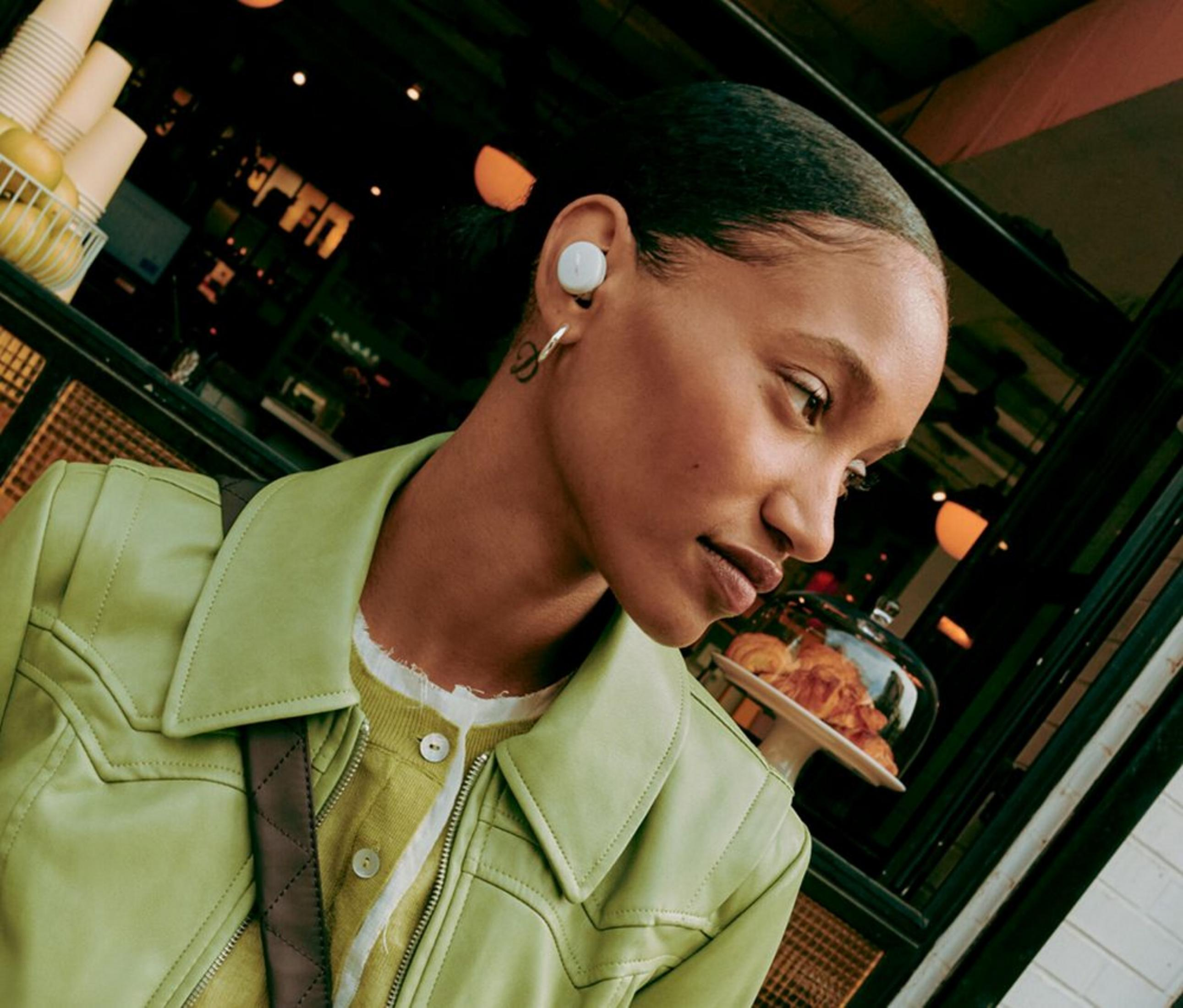 Una mujer joven lleva una chaqueta verde y auriculares blancos, mirando de lado en una cafetería.