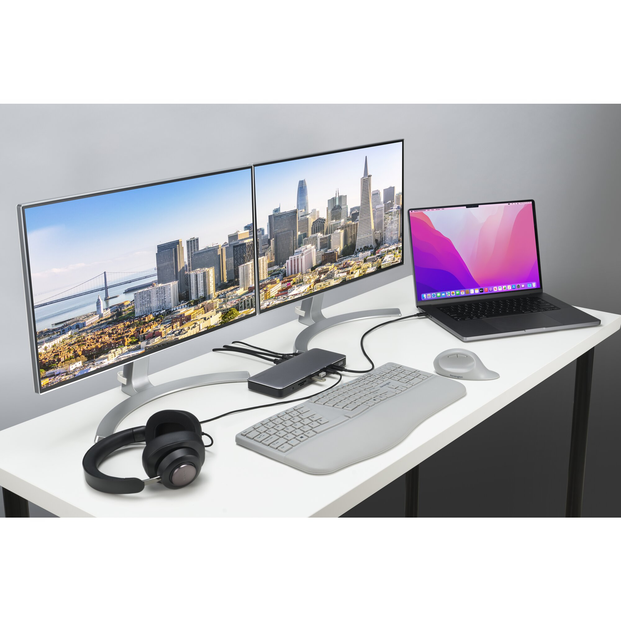 Desk with two monitors displaying city skyline, laptop, keyboard, and headphones.