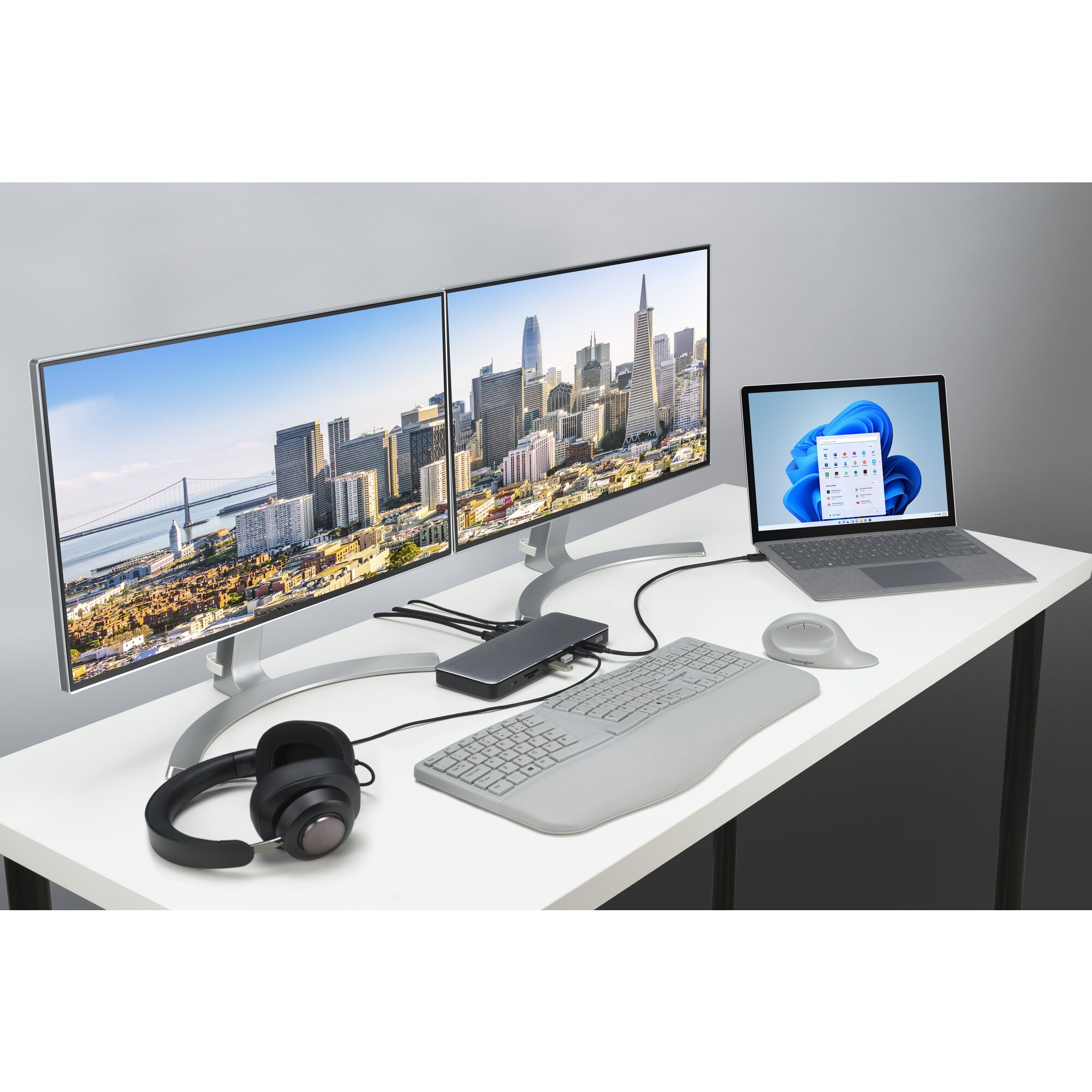 A desk setup with two monitors displaying a city skyline, laptop, keyboard, mouse, and headphones.