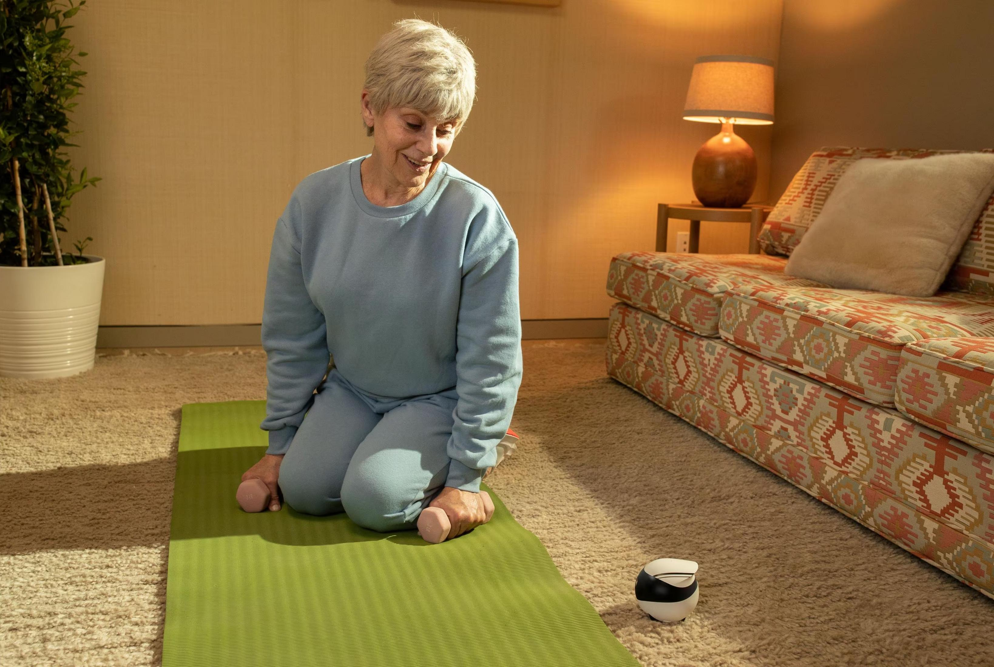 Starsza kobieta w niebieskim dresie klęczy na macie z hantlami.