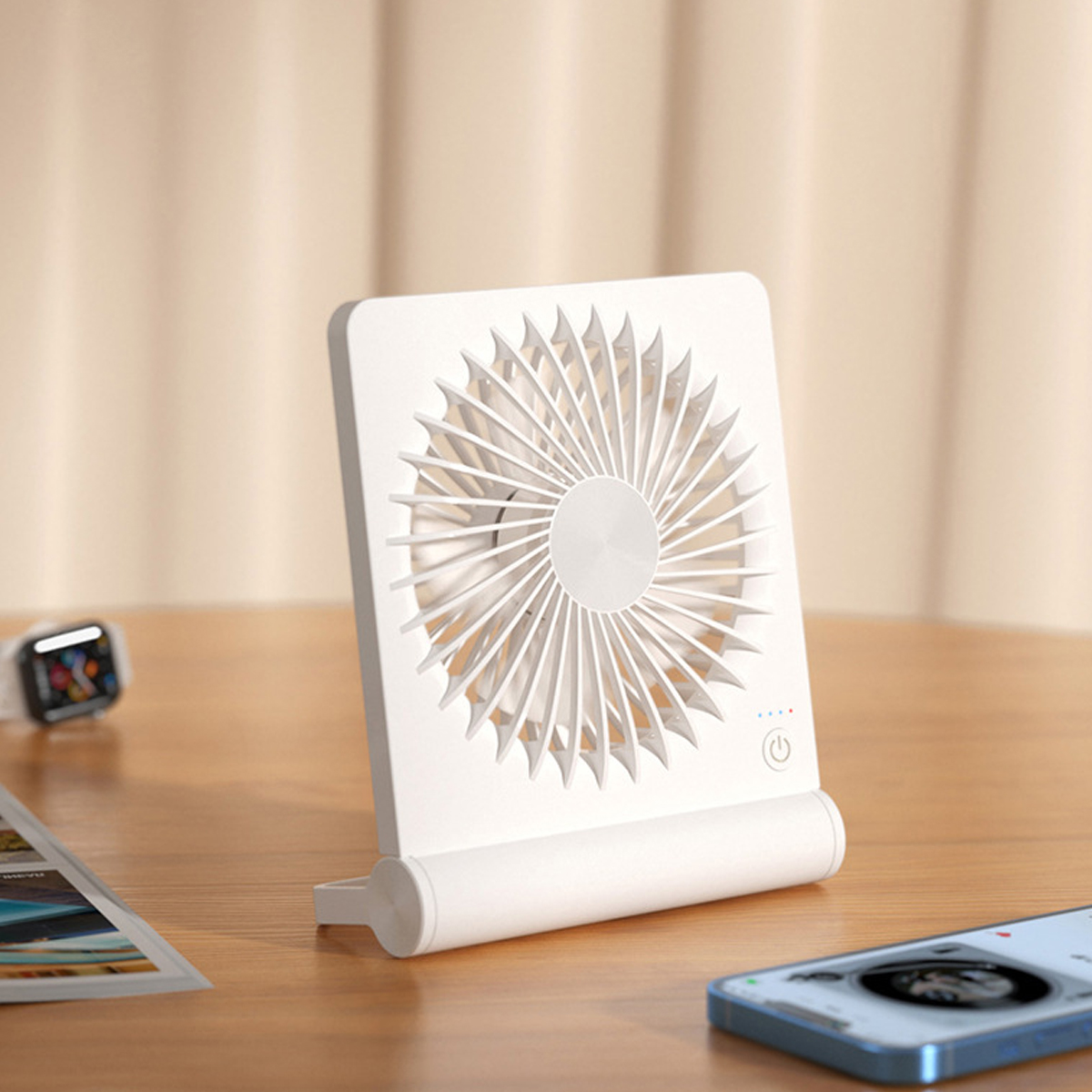 Ein weißer Tischventilator steht auf einem Holztisch, daneben ein Smartphone und Fotos.