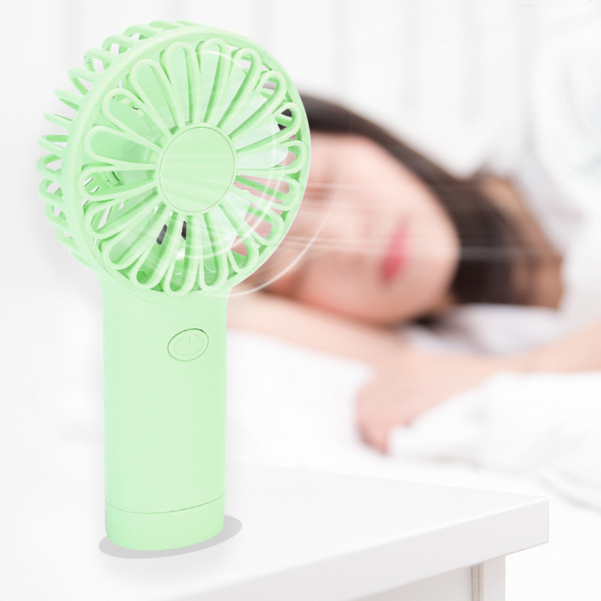 Ein hellgrüner Handventilator steht auf einem Tisch, eine Person schläft im Hintergrund.