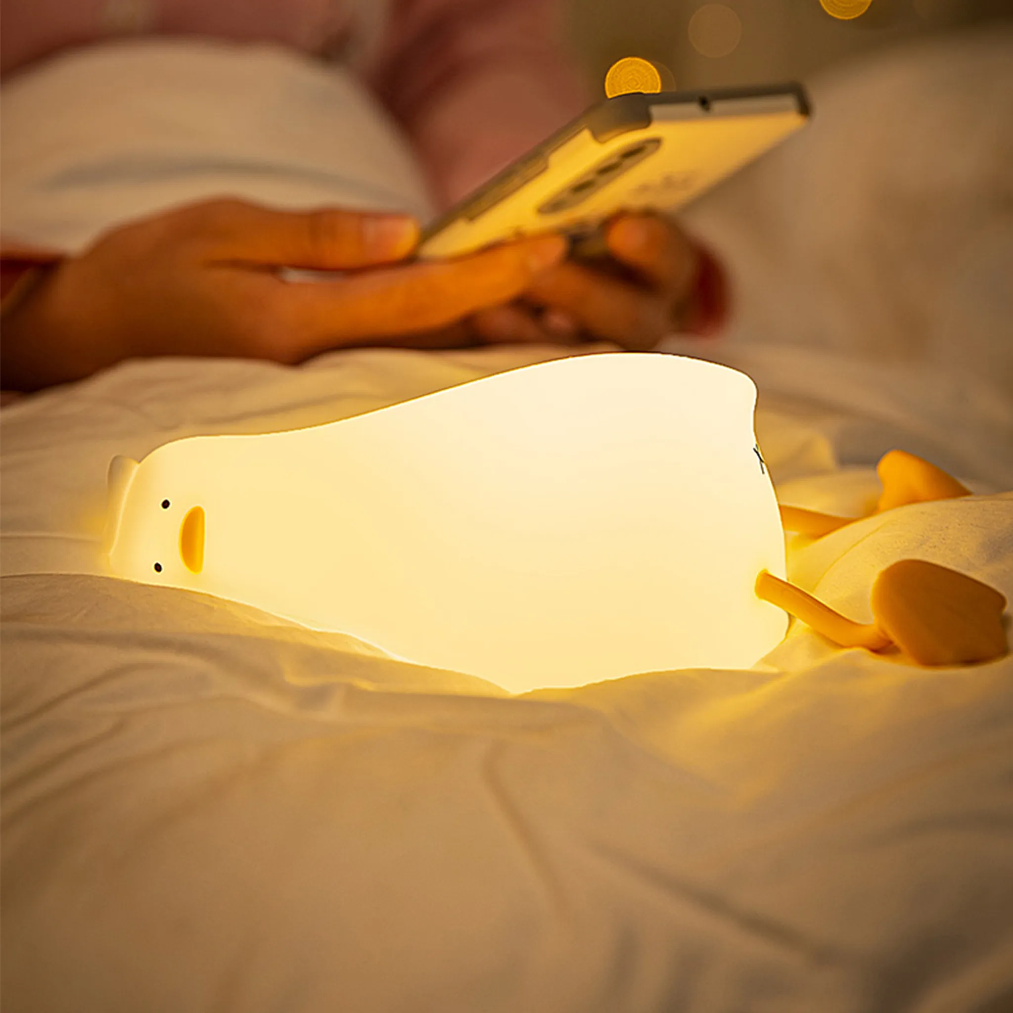 Eine leuchtende Ente auf einem Bett neben einer Person, die ein Smartphone benutzt.