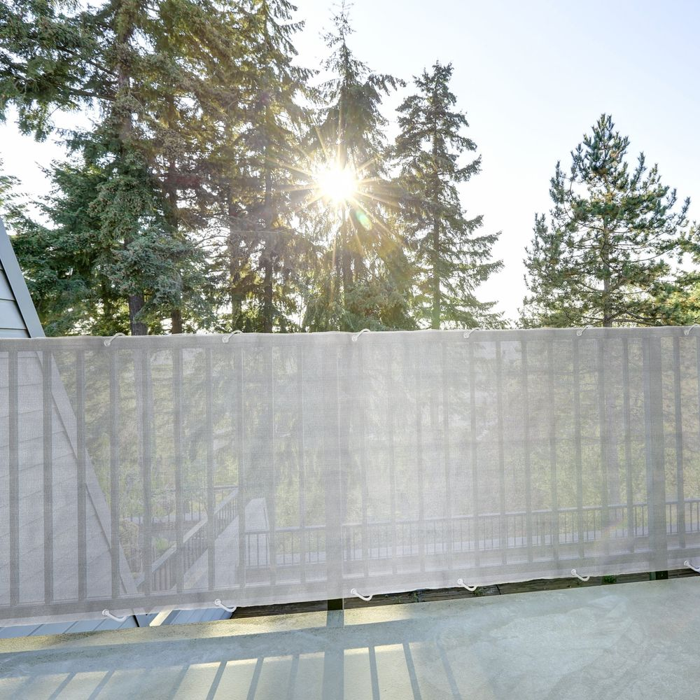 Eine helle, halbtransparente Sichtschutzrolle verdeckt eine Holzterrasse mit Bäumen im Hintergrund und Sonnenschein.