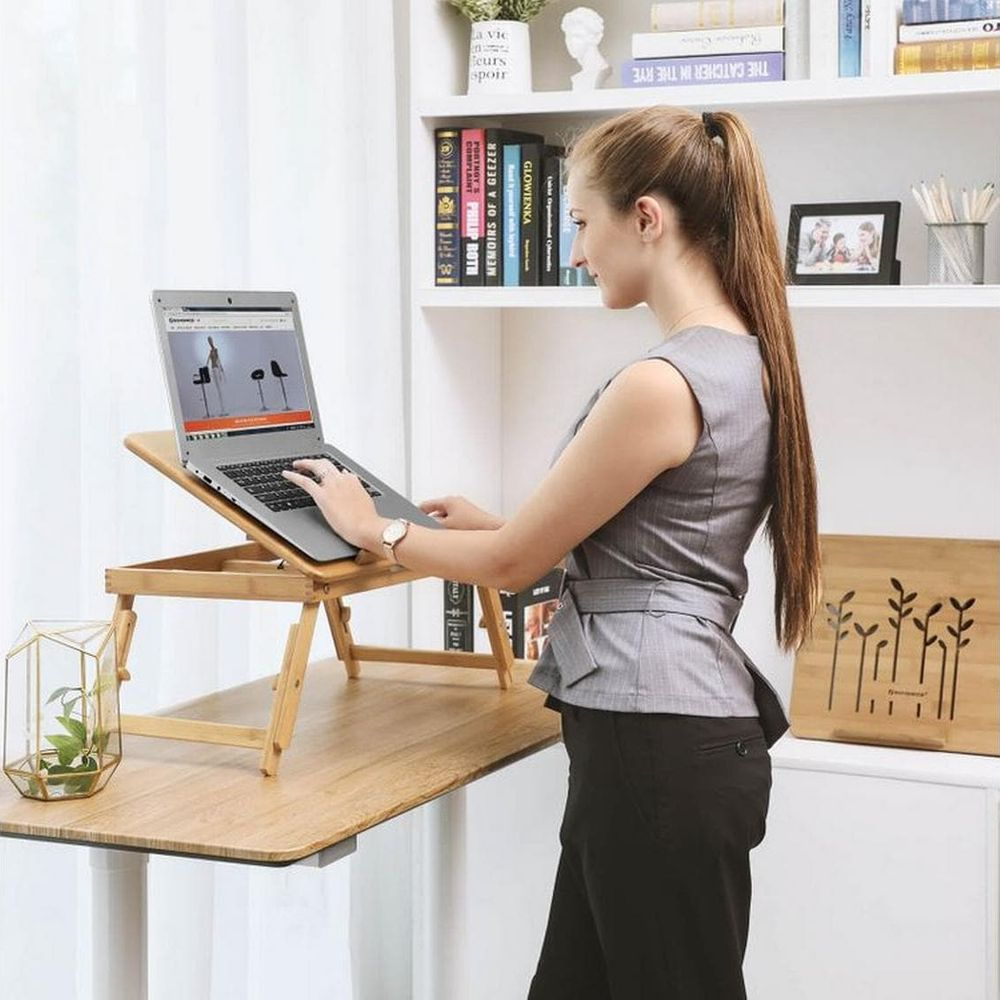 Frau steht und tippt auf einem Laptop auf einem Holztisch. Bücherregal im Hintergrund.