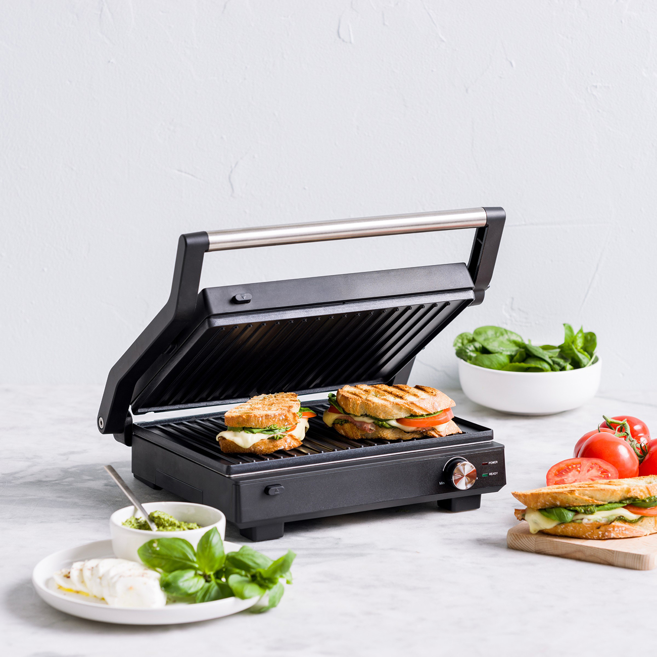 Ein offener Kontaktgrill mit zwei Sandwiches, Mozzarella, Basilikum und Tomaten.