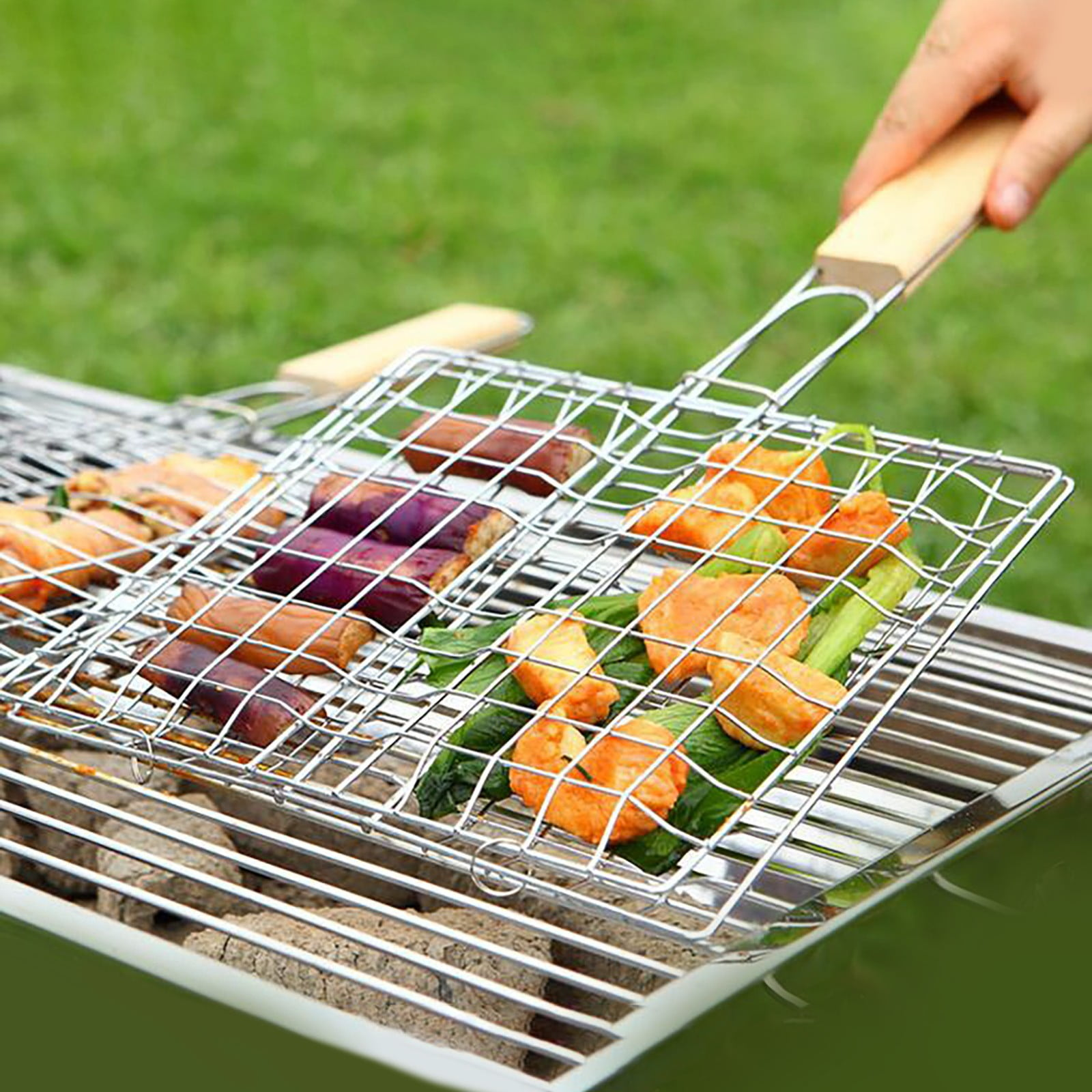 Ein Grillkorb mit Fleisch und Gemüse über glühenden Kohlen auf einer grünen Wiese.