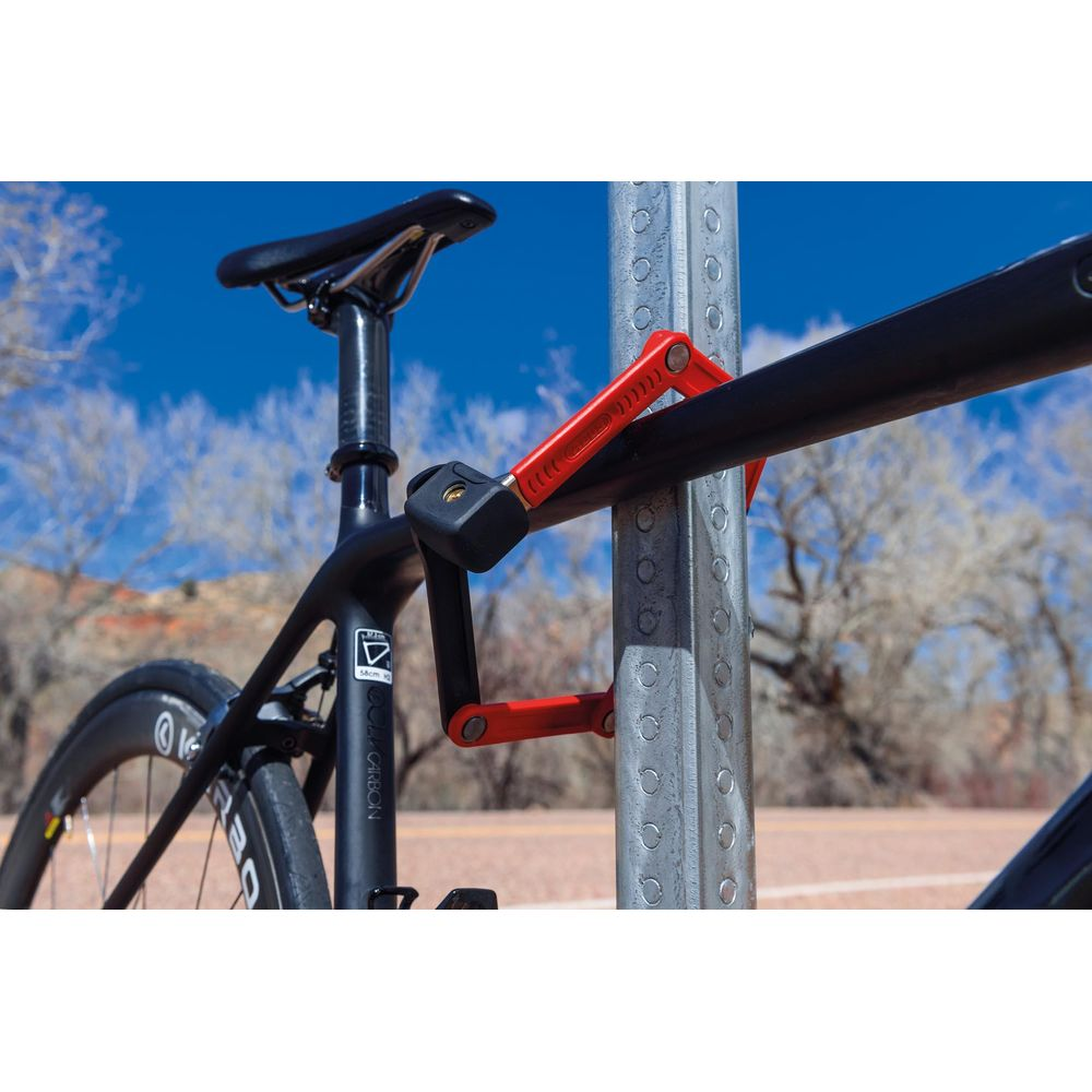 Ein rotes Faltschloss sichert ein schwarzes Fahrrad an einem Metallpfosten im Freien.