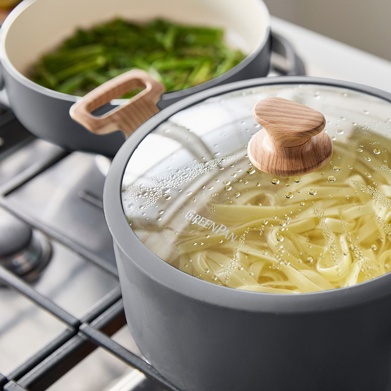 Fettuccine-Nudeln kochen in einem Topf mit Glasdeckel, daneben grüne Bohnen in einem Topf.