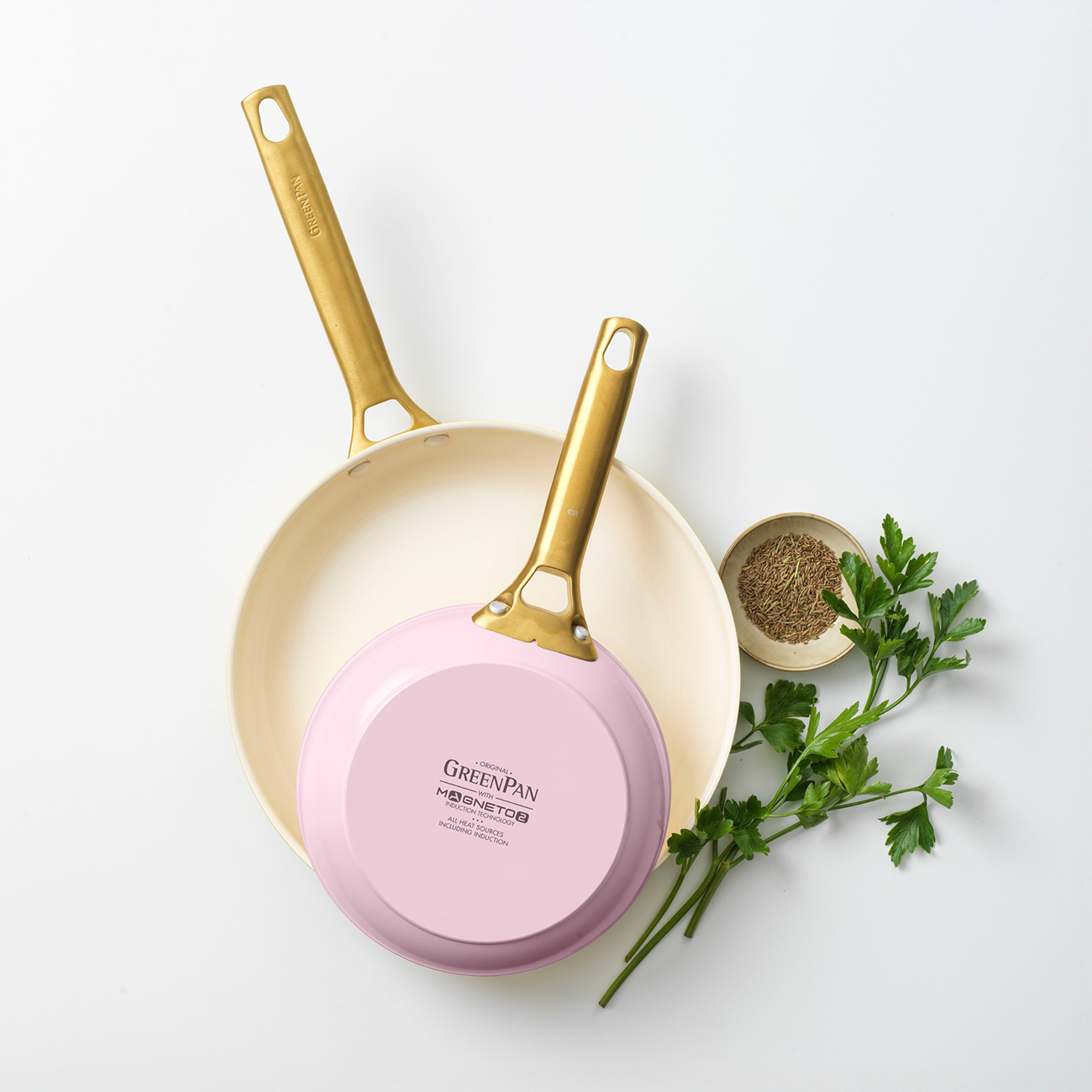 Zwei Pfannen mit goldenen Griffen, eine rosa, eine cremefarben, mit Kräutern und Samen.