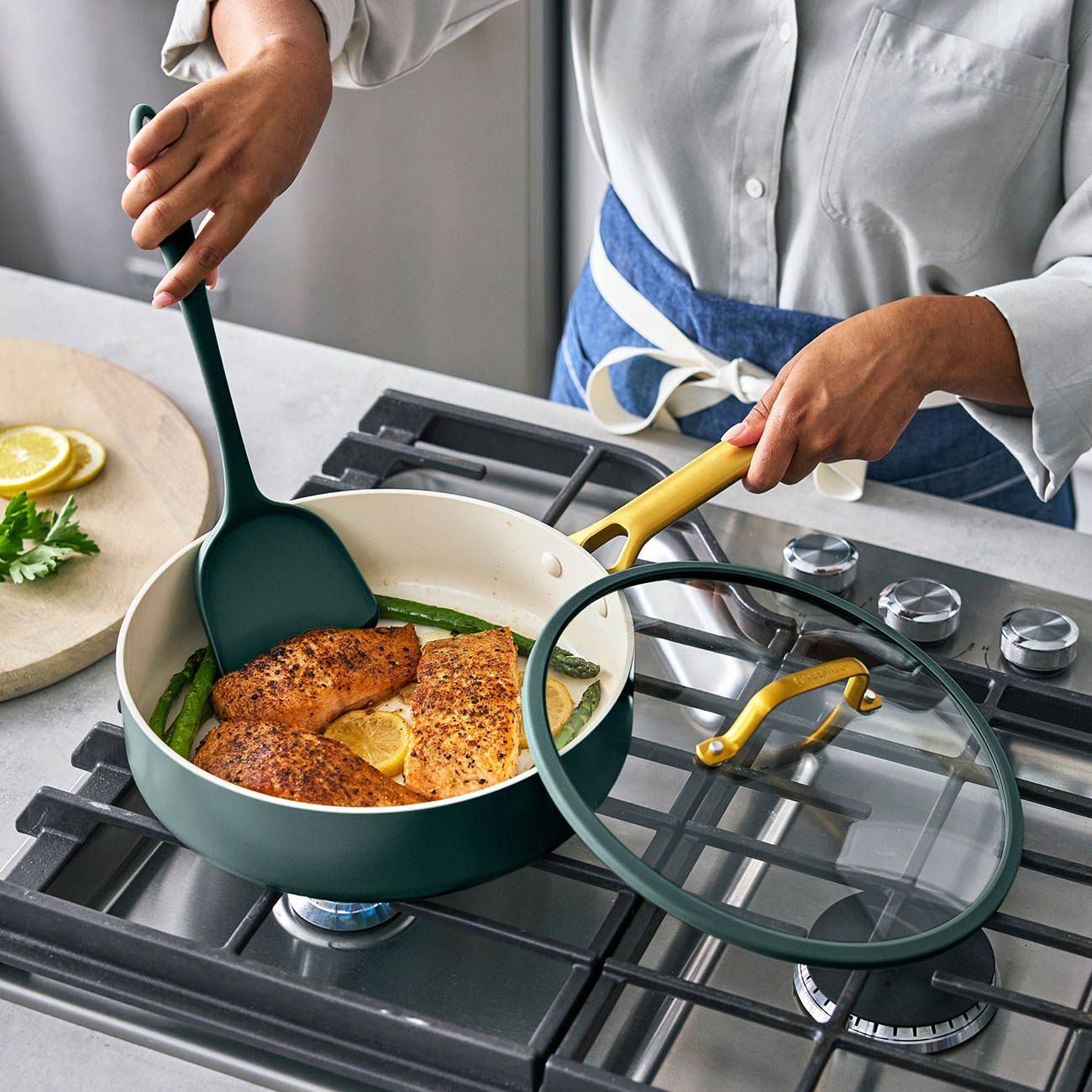 Person kocht Lachs mit Spargel und Zitrone in einer grünen Pfanne.