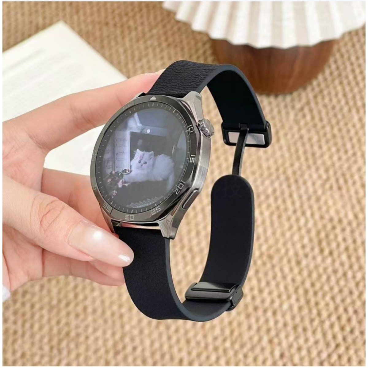 Una mano tiene uno smartwatch argentato con cinturino nero. Il display mostra un gatto bianco.
