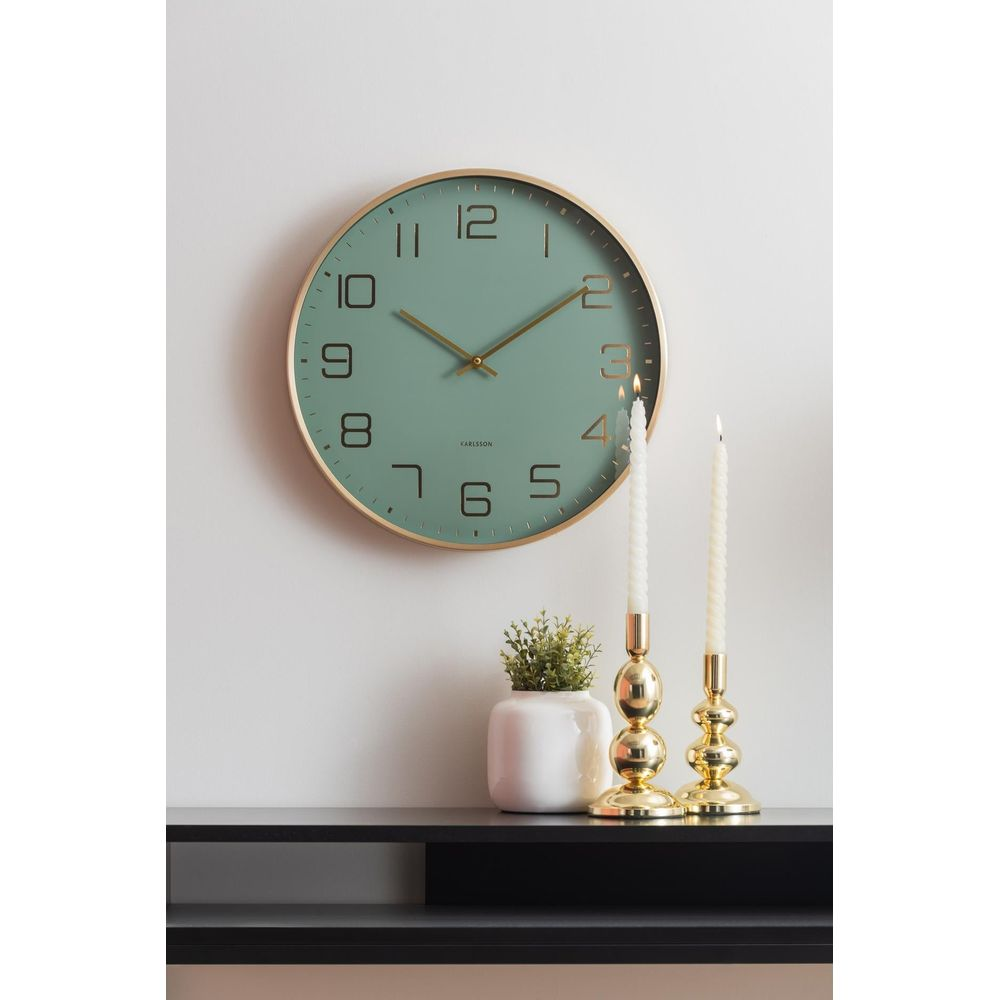 Grüne Wanduhr mit goldenem Rahmen, daneben zwei Kerzenhalter mit Kerzen und eine kleine Pflanze.
