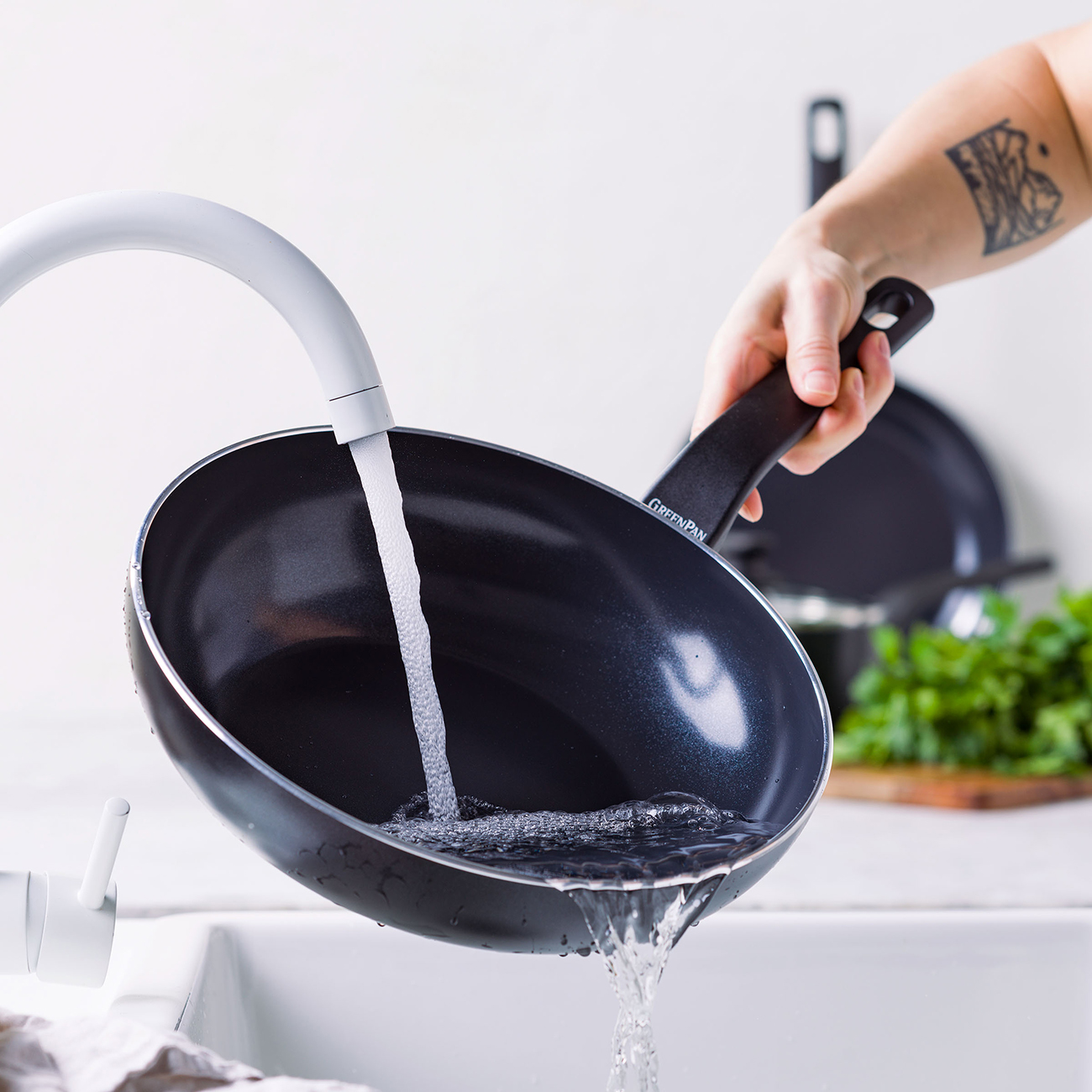 Eine Hand hält eine schwarze Pfanne, während Wasser aus dem Hahn hineinfließt.