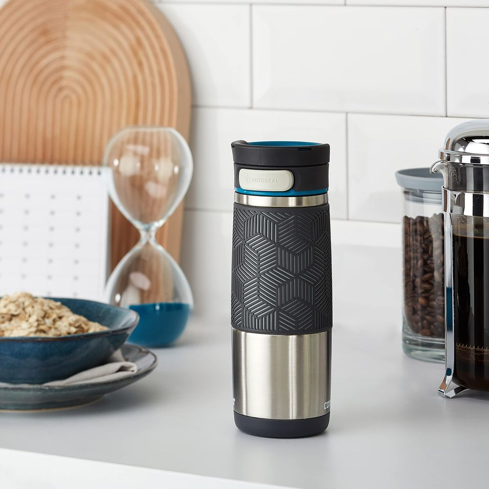 Edelstahlbecher mit schwarzem Griff auf einer Arbeitsplatte mit Haferflocken und Kaffeemaschine.