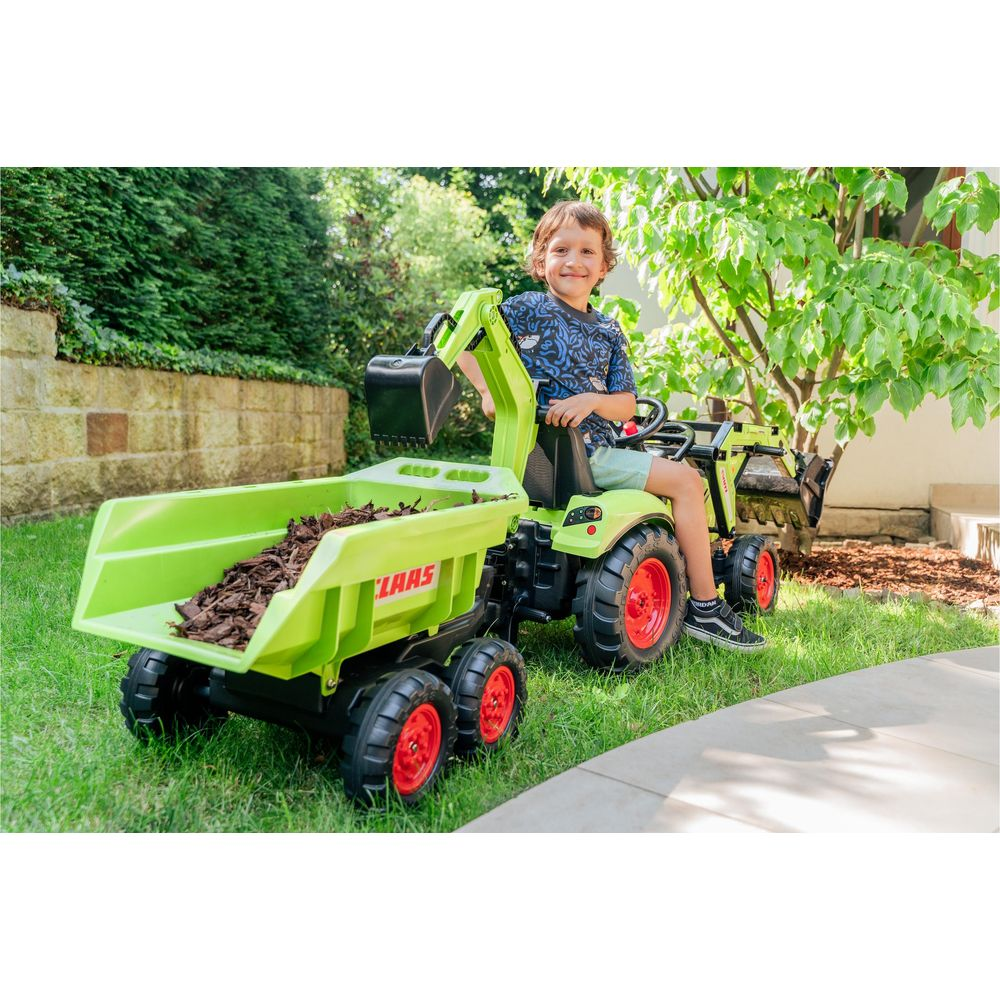 Ein Junge sitzt auf einem grünen Spielzeugtraktor mit Anhänger und Schaufel.
