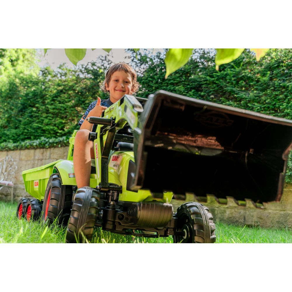 Ein Junge sitzt auf einem grünen Spielzeugtraktor mit Schaufel und Anhänger im Garten.