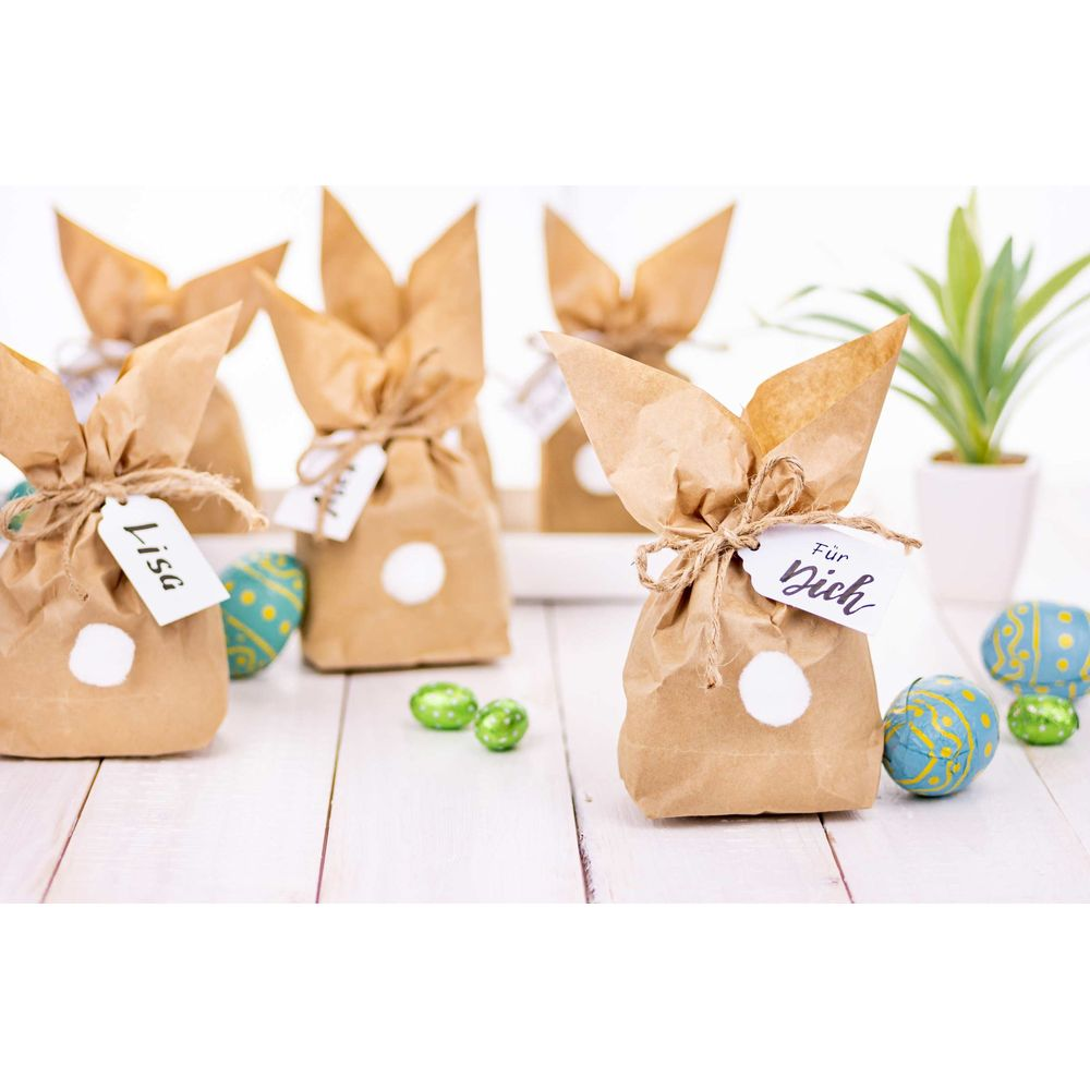 Sacchetti regalo marroni a forma di coniglio con cerchi bianchi e etichette, circondati da uova di Pasqua.