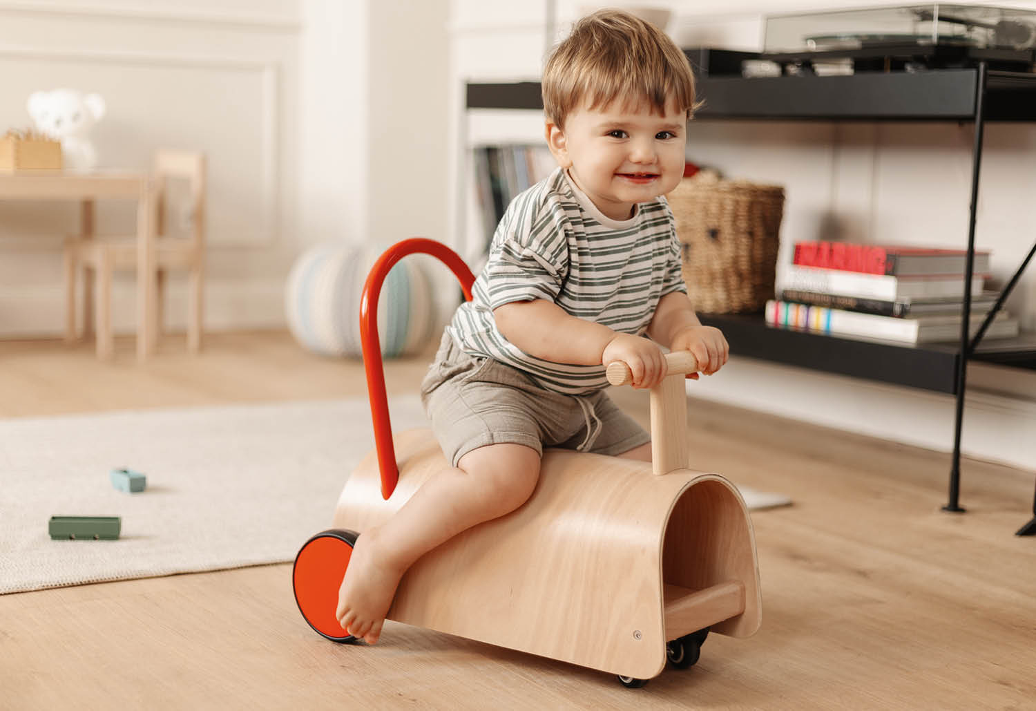 Ein Baby sitzt auf einem hölzernen Spielzeugauto mit rotem Griff und lächelt.