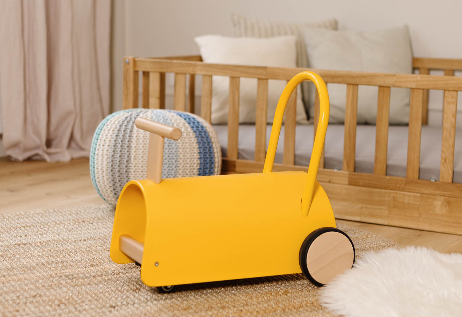 Ein gelbes Lauflernspielzeug mit Holzgriffen und Rädern steht auf einem Teppich neben einem Kinderbett.