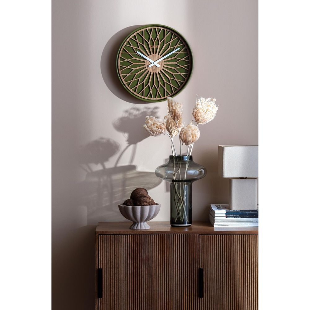 Un orologio decorativo è appeso al muro sopra un mobile in legno con un vaso di fiori secchi.