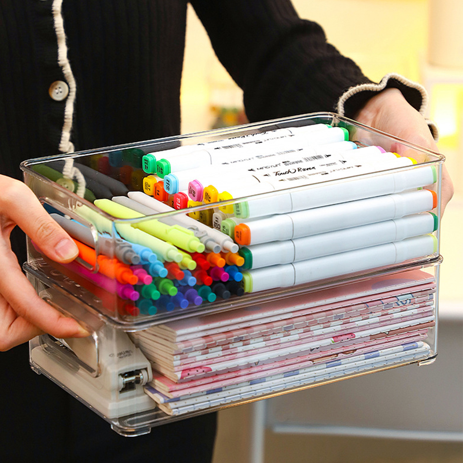 Transparenter Plastikorganizer mit bunten Markern oben und gestapelten Notizbüchern unten.