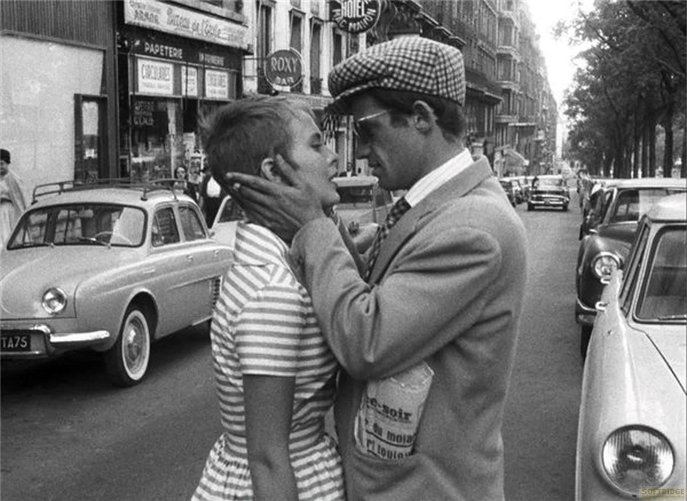 Un uomo con cappello bacia una donna in strada tra auto e negozi.