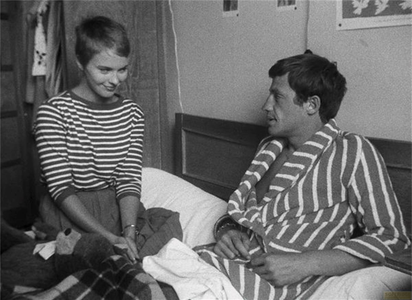 Un uomo e una donna seduti su un letto. Lei indossa un top a righe. Lui indossa una vestaglia.