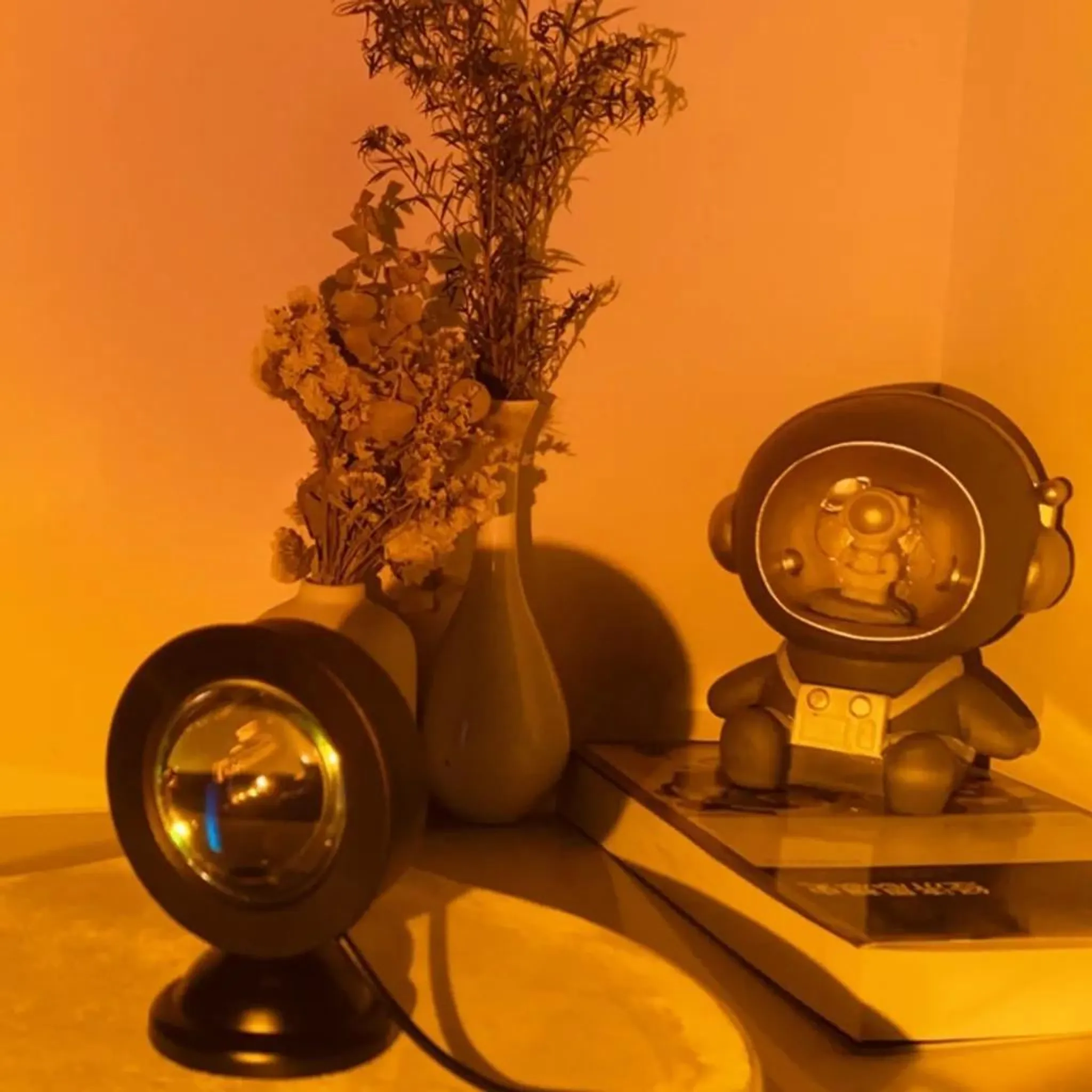 Eine Astronautenfigur sitzt auf Büchern neben Vasen mit Trockenblumen und einer Sonnenuntergangslampe.