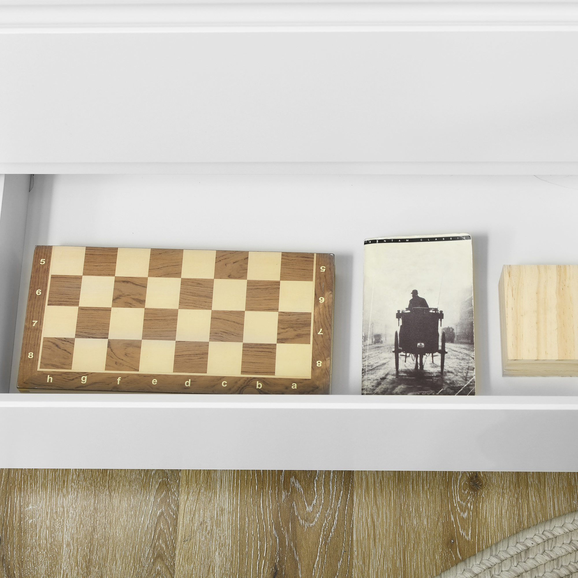 Un tablero de ajedrez, un libro con un carruaje y un bloque de madera descansan en un estante blanco.