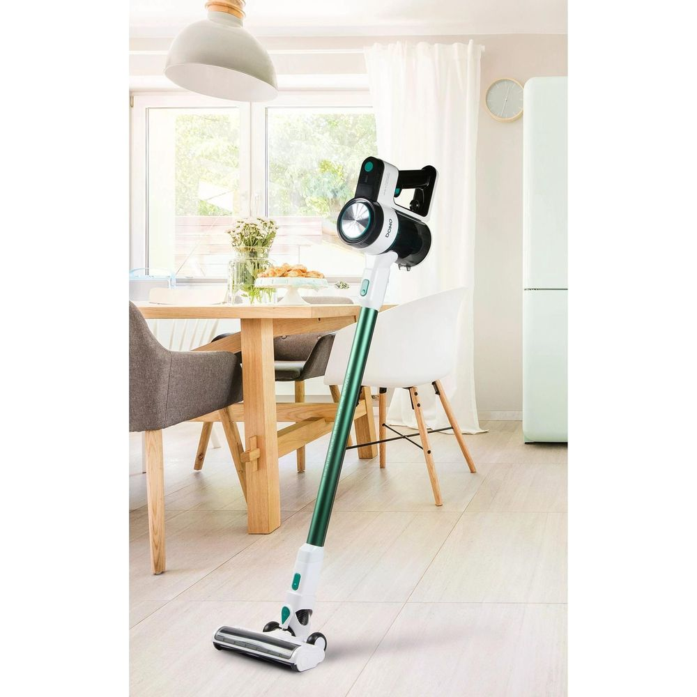 Un aspirateur sans fil vert et blanc se tient dans une cuisine avec table et chaises en bois.