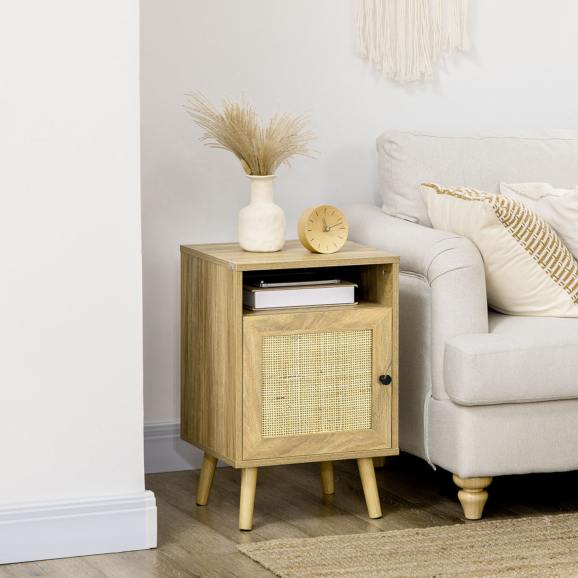 Mesita de noche de madera con puerta de ratán, jarrón con flores secas, reloj y sofá.