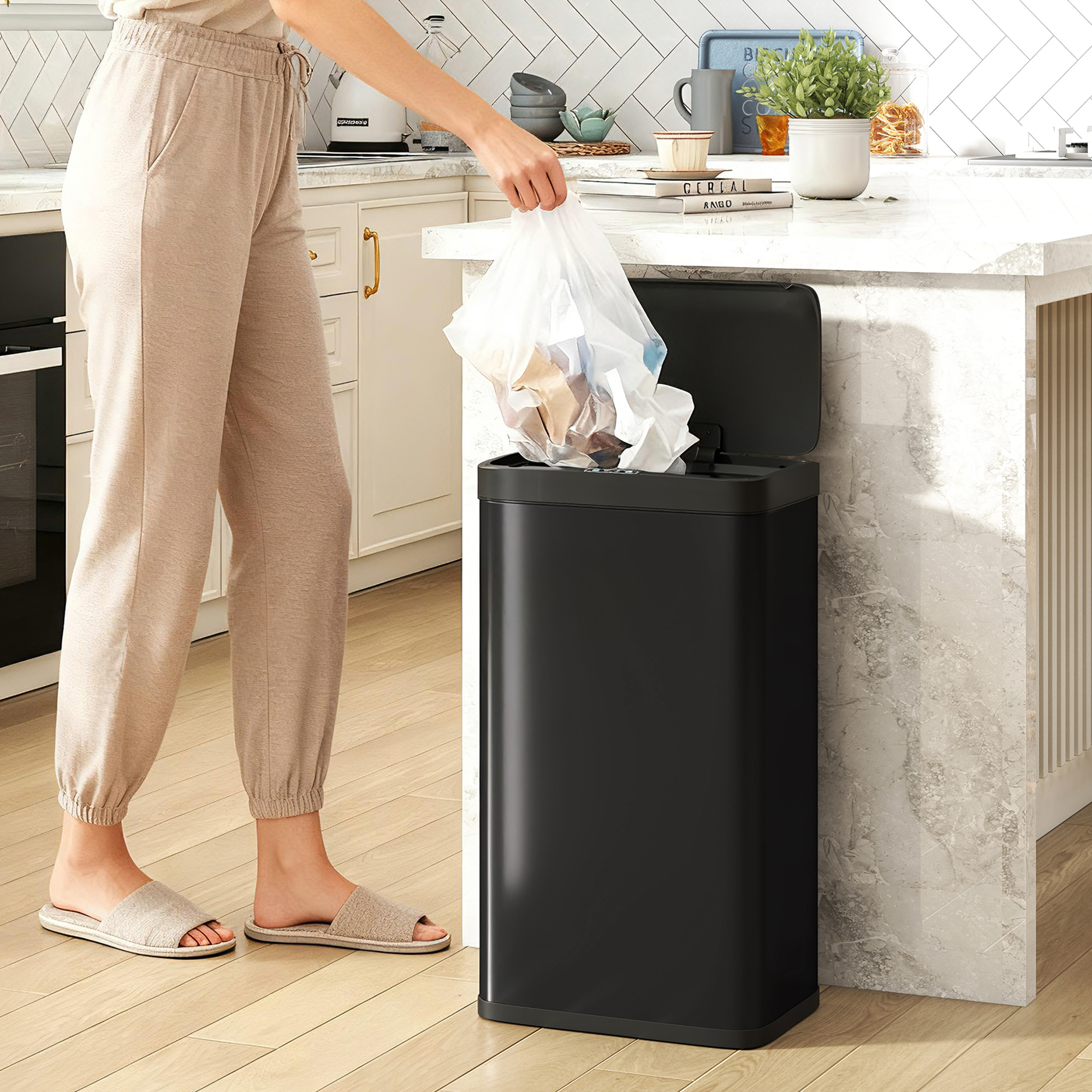 Una persona con pantalones beige y pantuflas tira basura en un cubo negro en una cocina moderna.