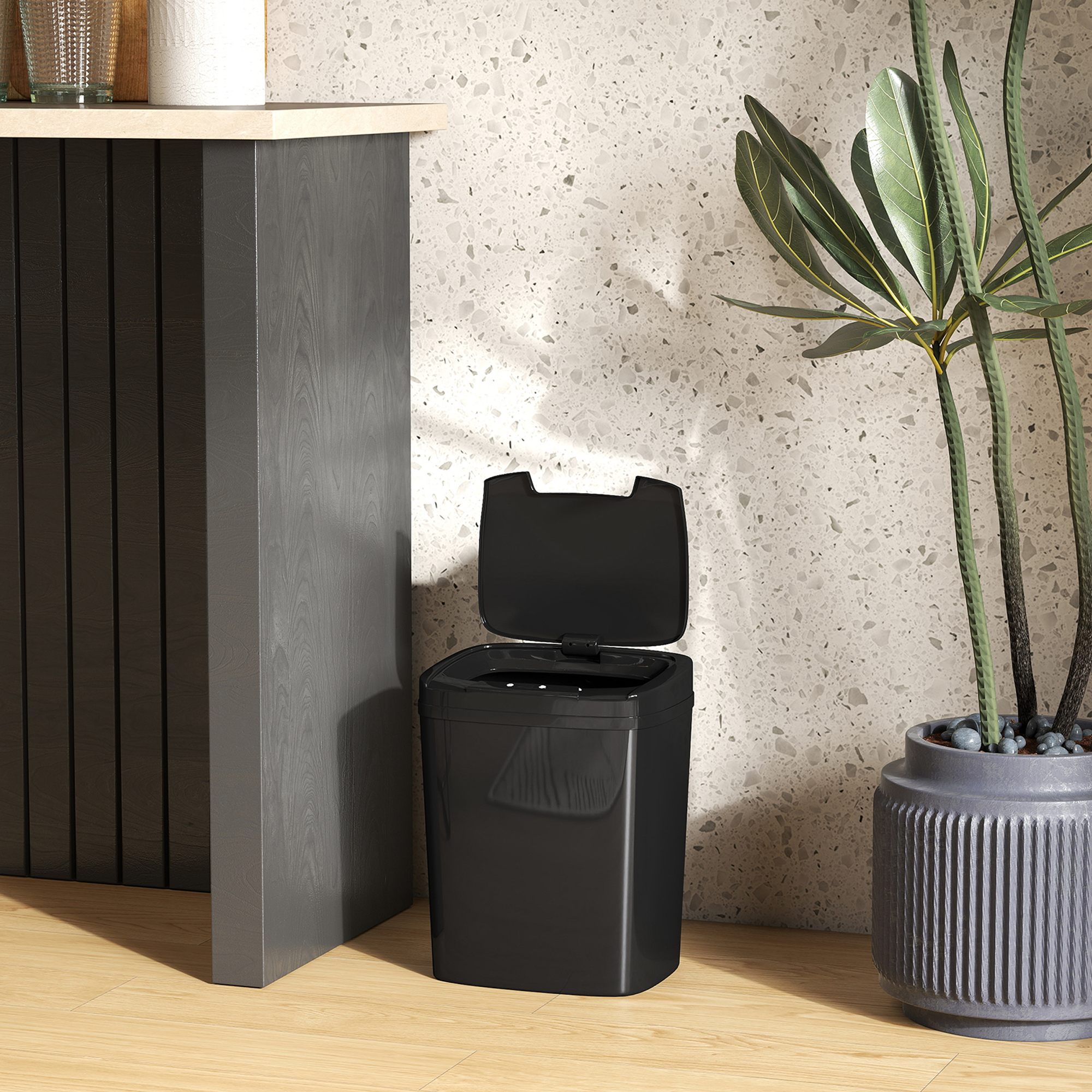 Un cubo de basura negro con tapa abierta junto a una planta verde en maceta.