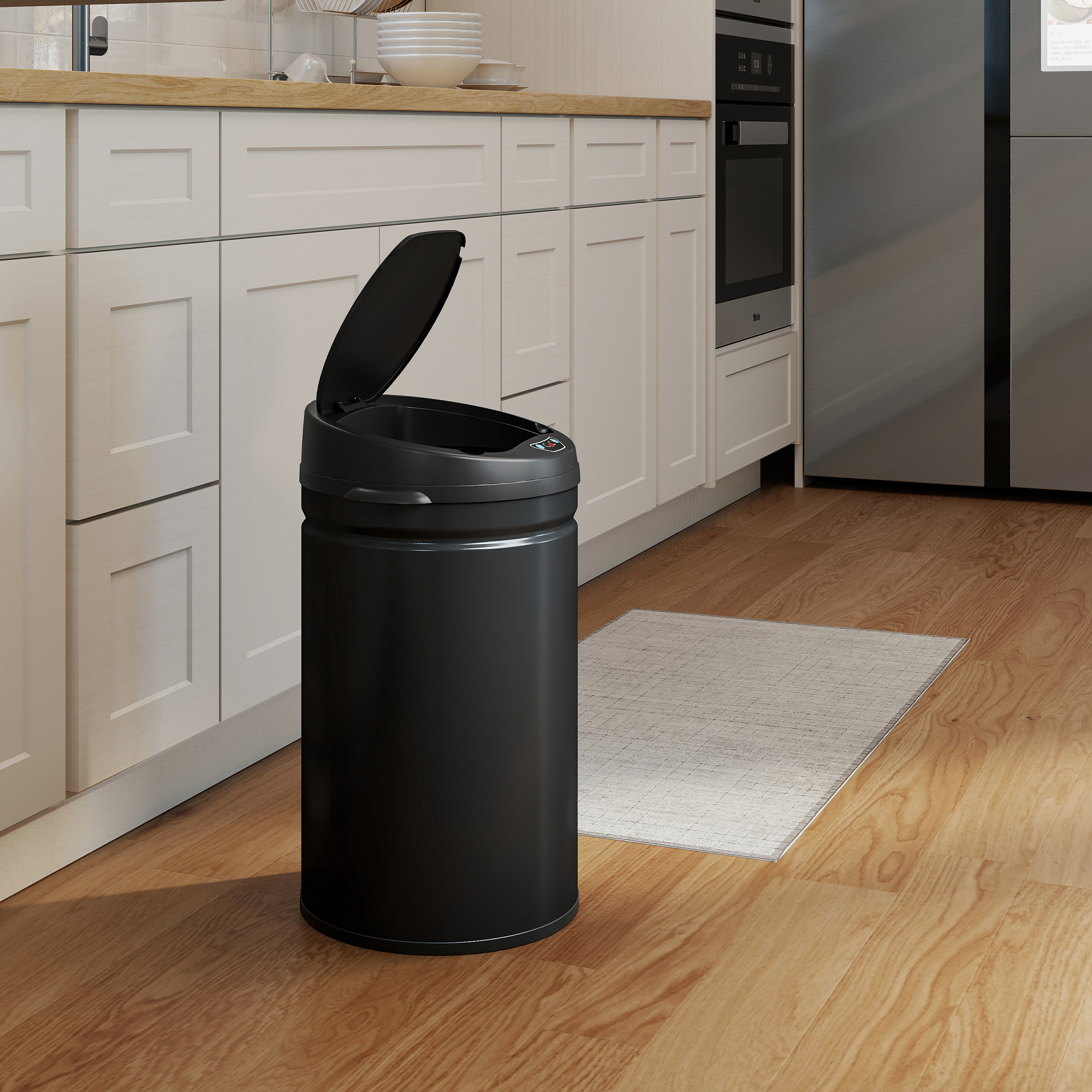 Un cubo de basura negro con tapa abierta sobre suelo de madera junto a armarios blancos.