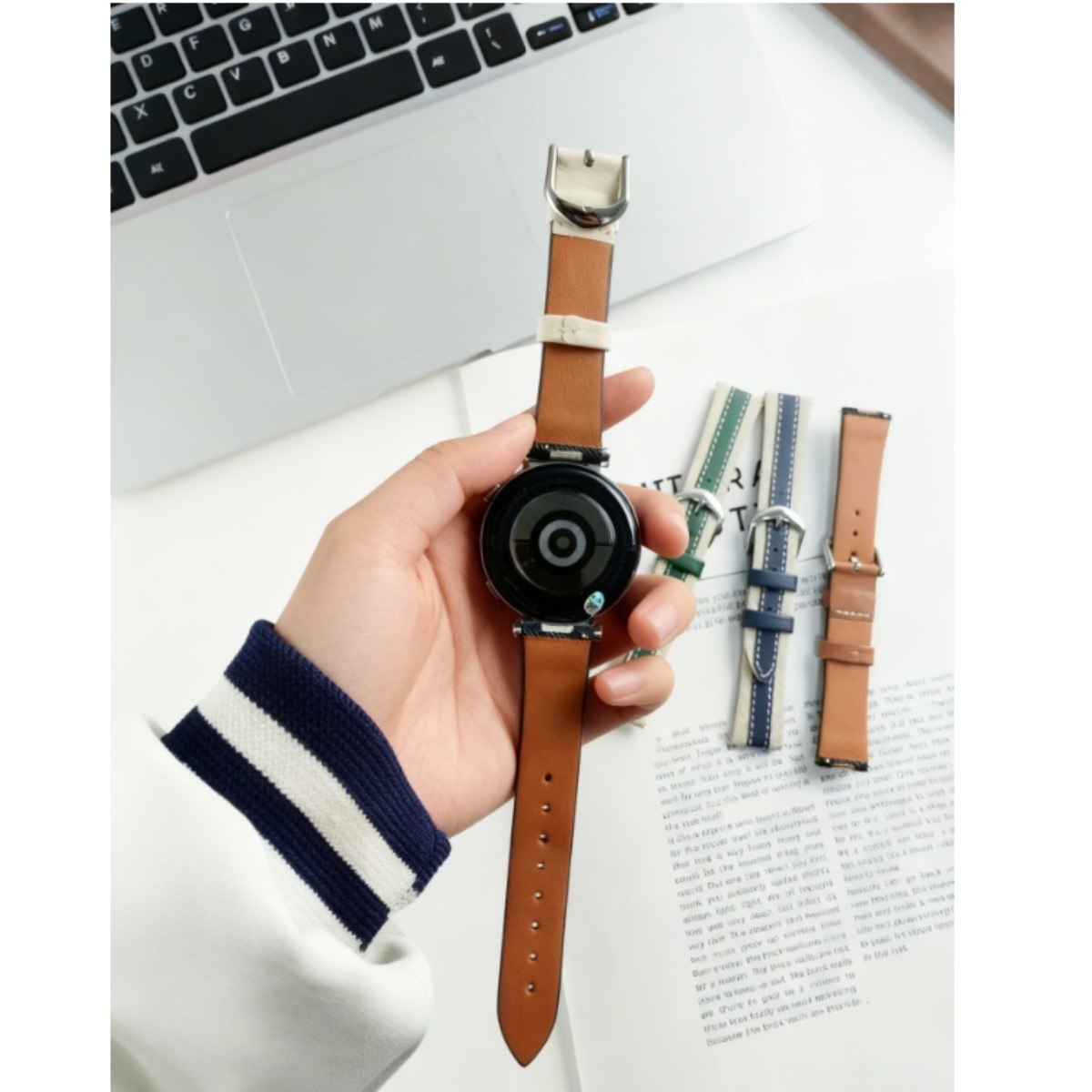 Eine Hand hält eine Smartwatch mit braunem Lederarmband. Mehrere Armbänder liegen daneben.