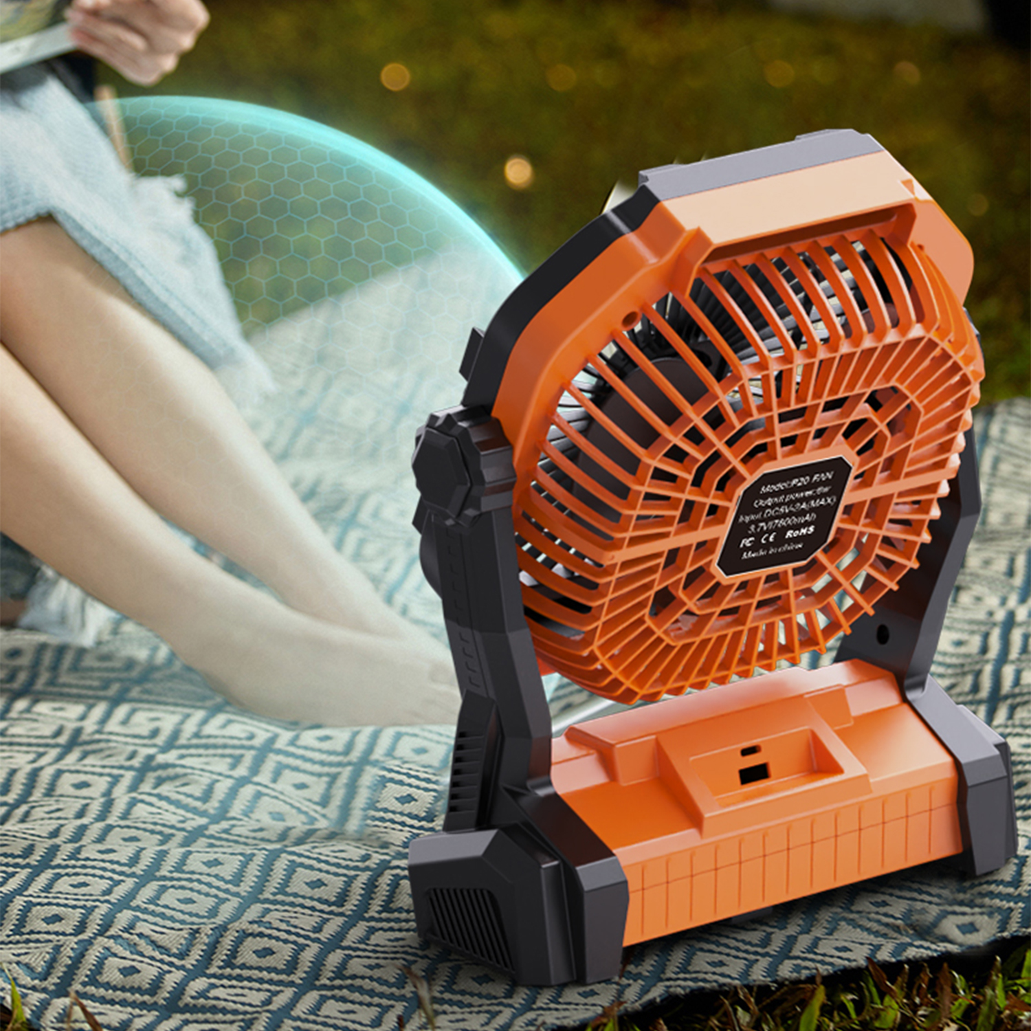 Un ventilatore portatile arancione è su una coperta accanto alle gambe di una persona seduta.