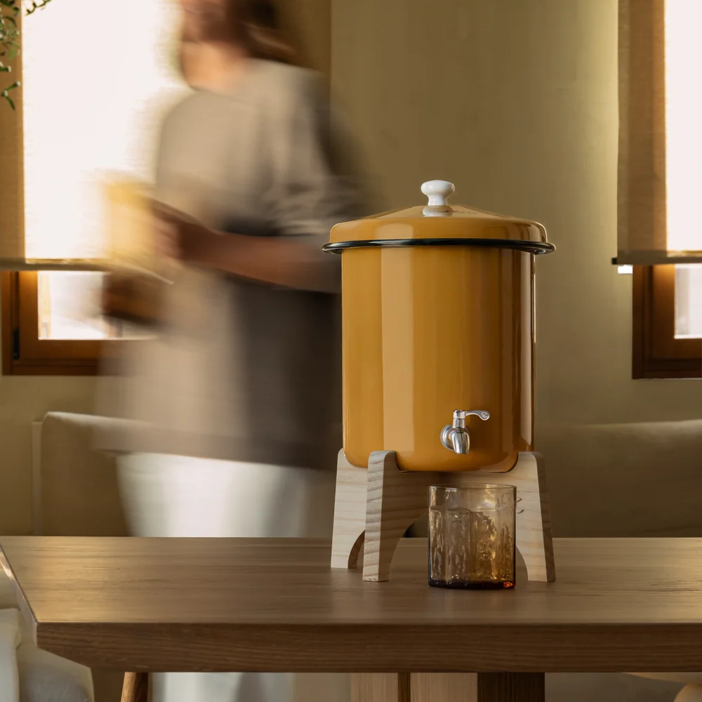 Un erogatore giallo senape con rubinetto poggia su un supporto di legno accanto a un bicchiere.