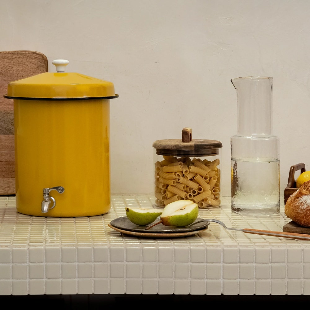 Un dispenser giallo con rubinetto, pasta in barattolo, due metà di pera e una brocca d'acqua.