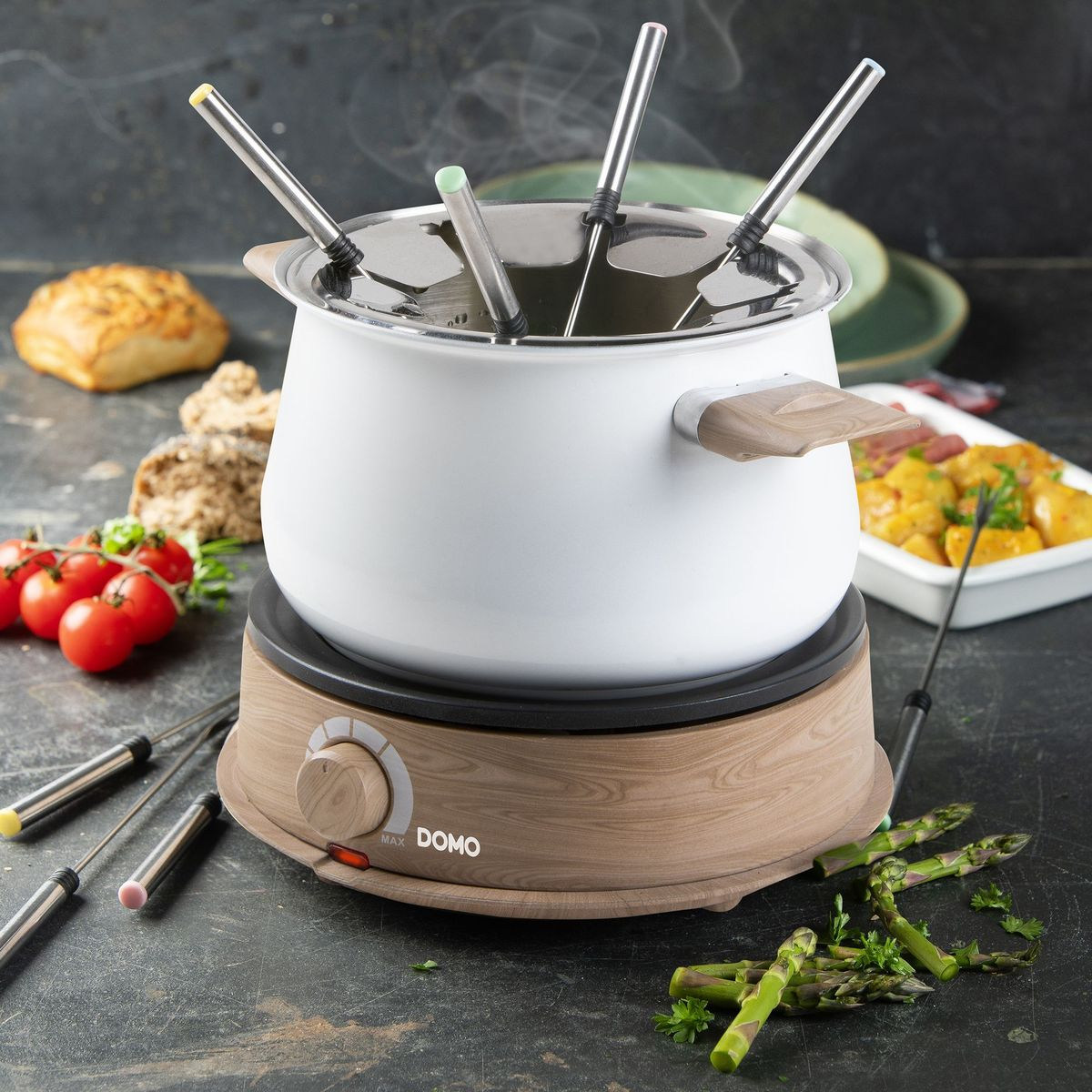 Ein weißer Fondue-Topf mit Holzsockel, umgeben von Zutaten wie Tomaten, Brot und Spargel.