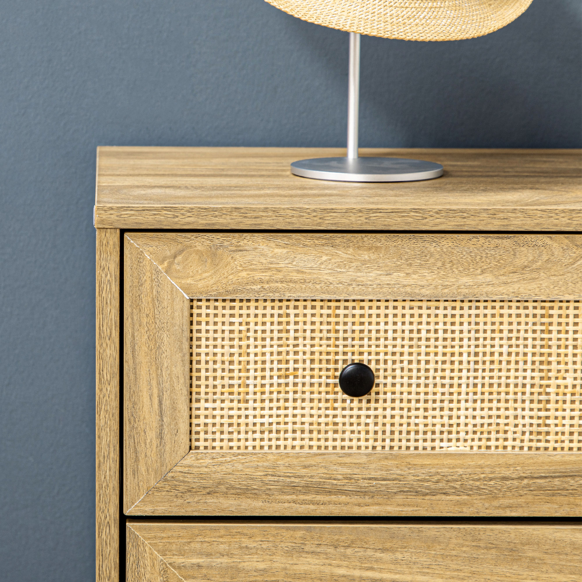 Cómoda de madera con frente de ratán y pomo negro sobre pared azul.