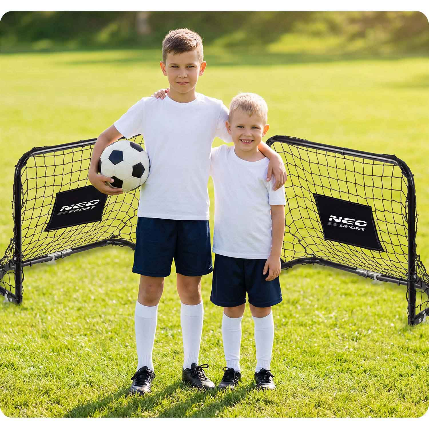 Dwóch chłopców w strojach piłkarskich stoi obok małych bramek na trawiastym boisku.
