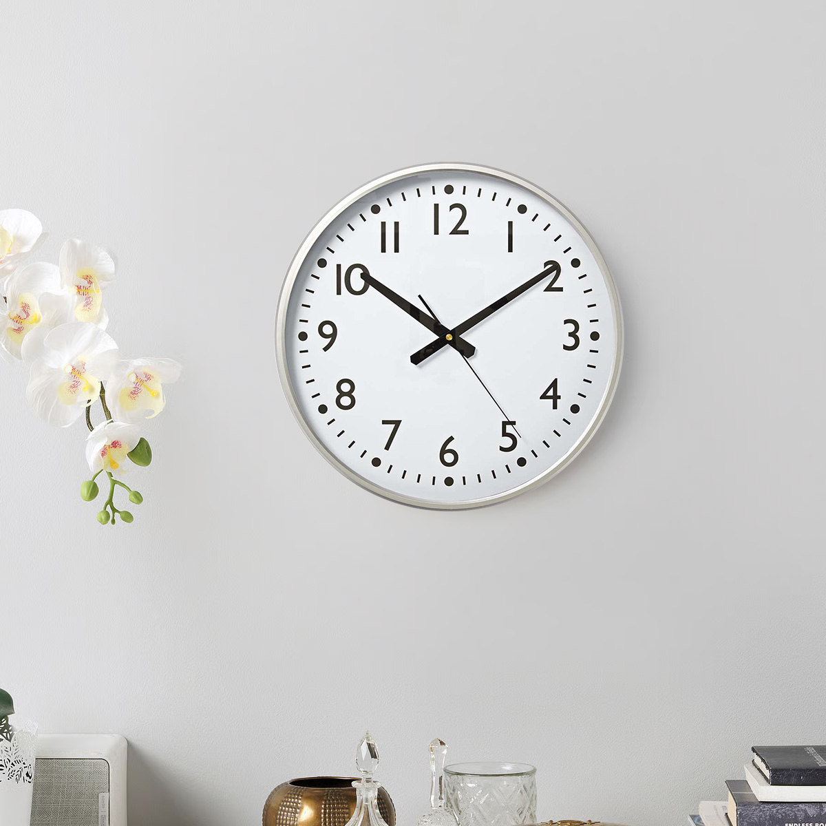 Eine weiße Wanduhr mit schwarzen Zahlen und Zeigern hängt an einer grauen Wand.