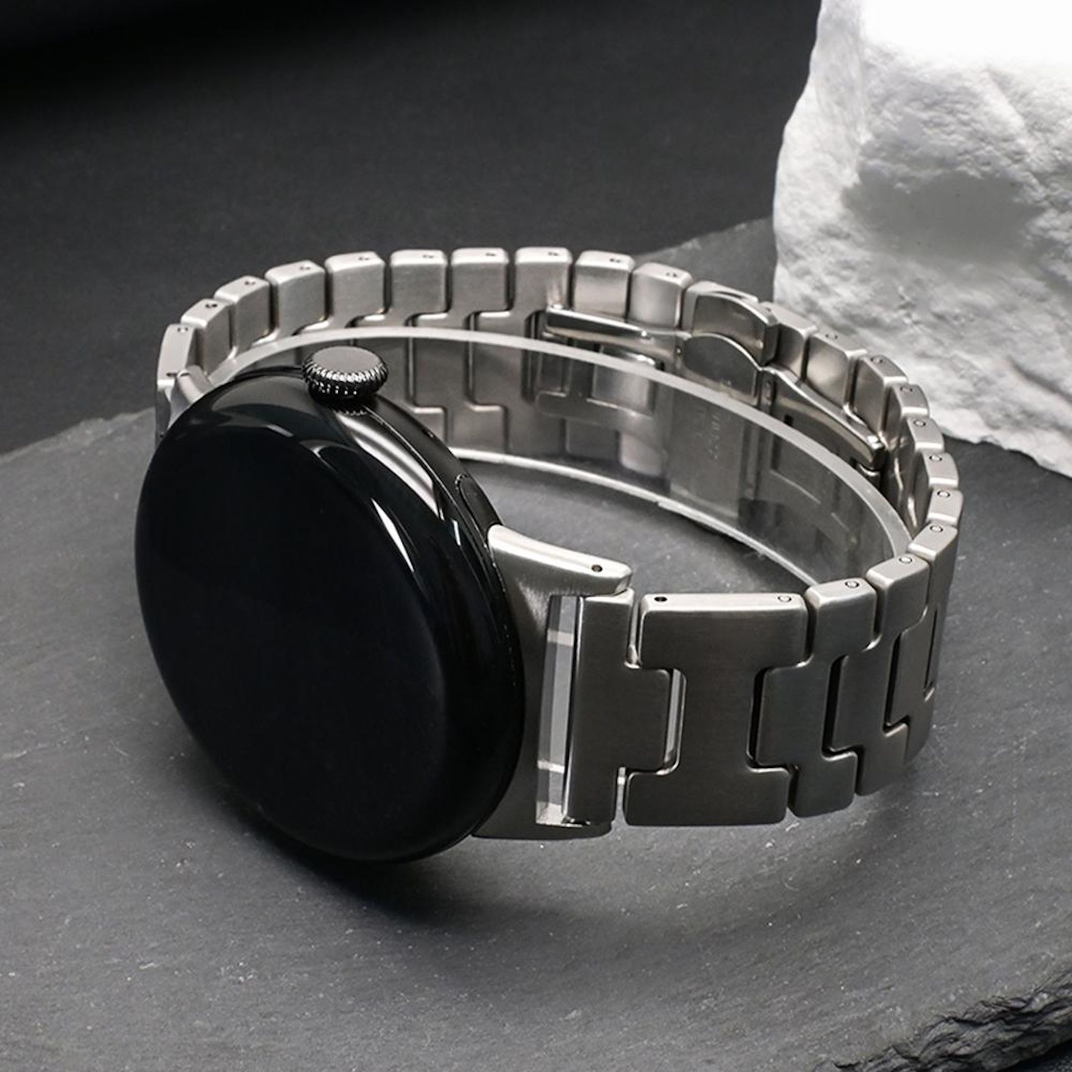 Uno smartwatch nero con cinturino in metallo argentato riposa su pietra scura.