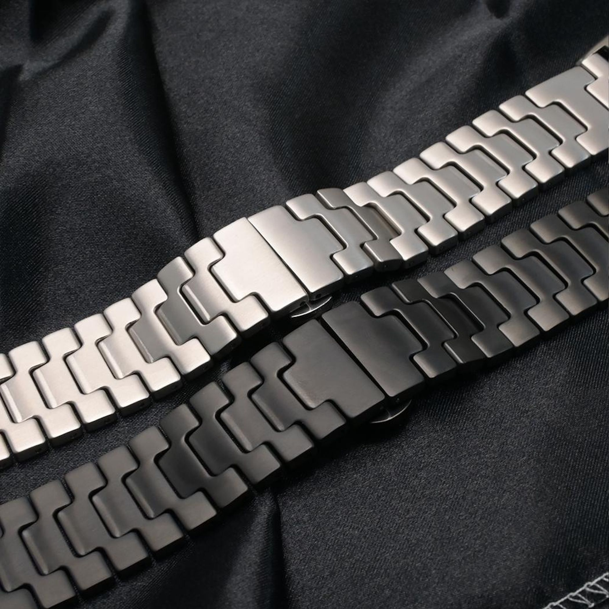 Zwei Edelstahl-Uhrenarmbänder, eines silbern, eines schwarz, liegen auf schwarzem Stoff.