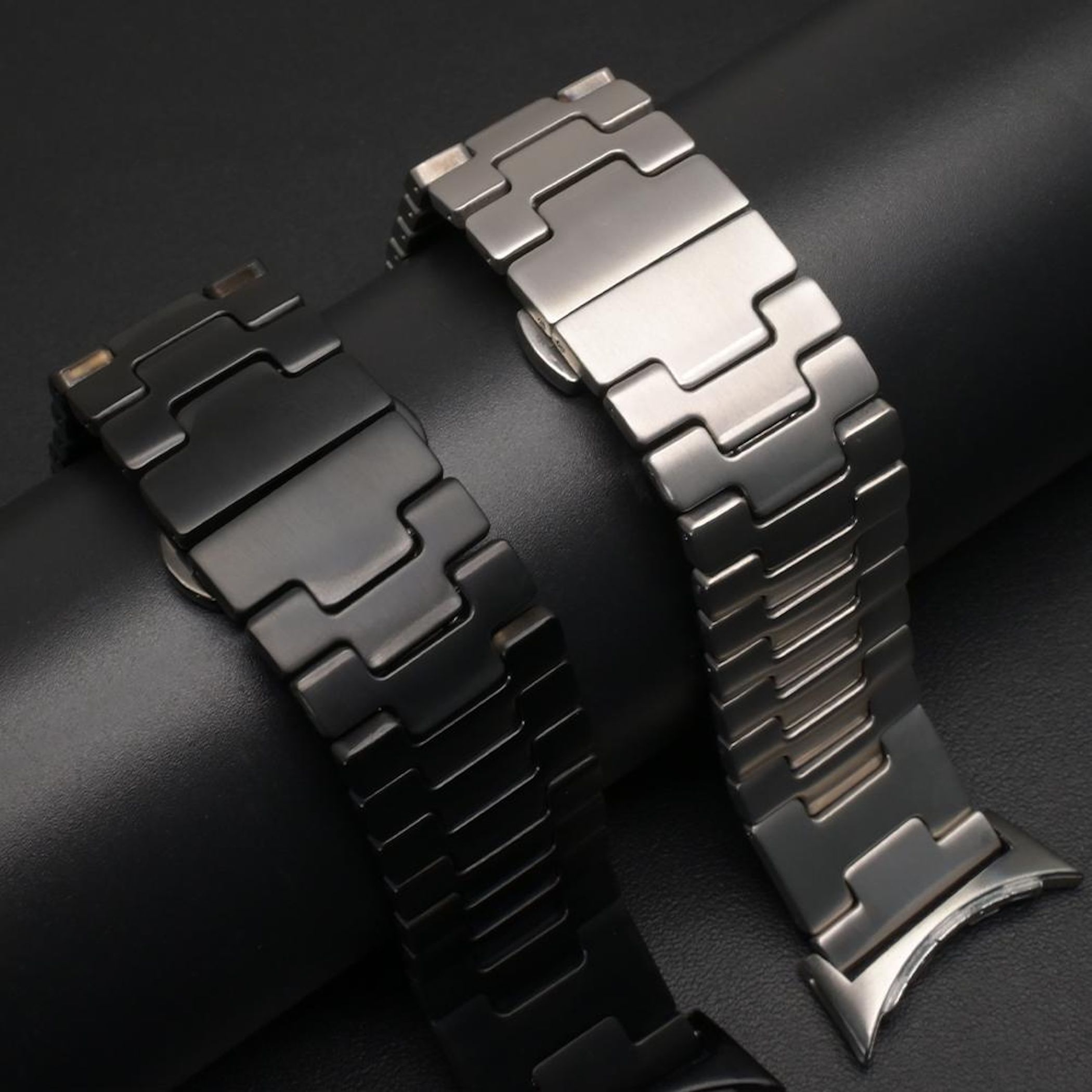 Zwei metallische Uhrenarmbänder, eines schwarz, eines silbern, auf schwarzem Hintergrund.